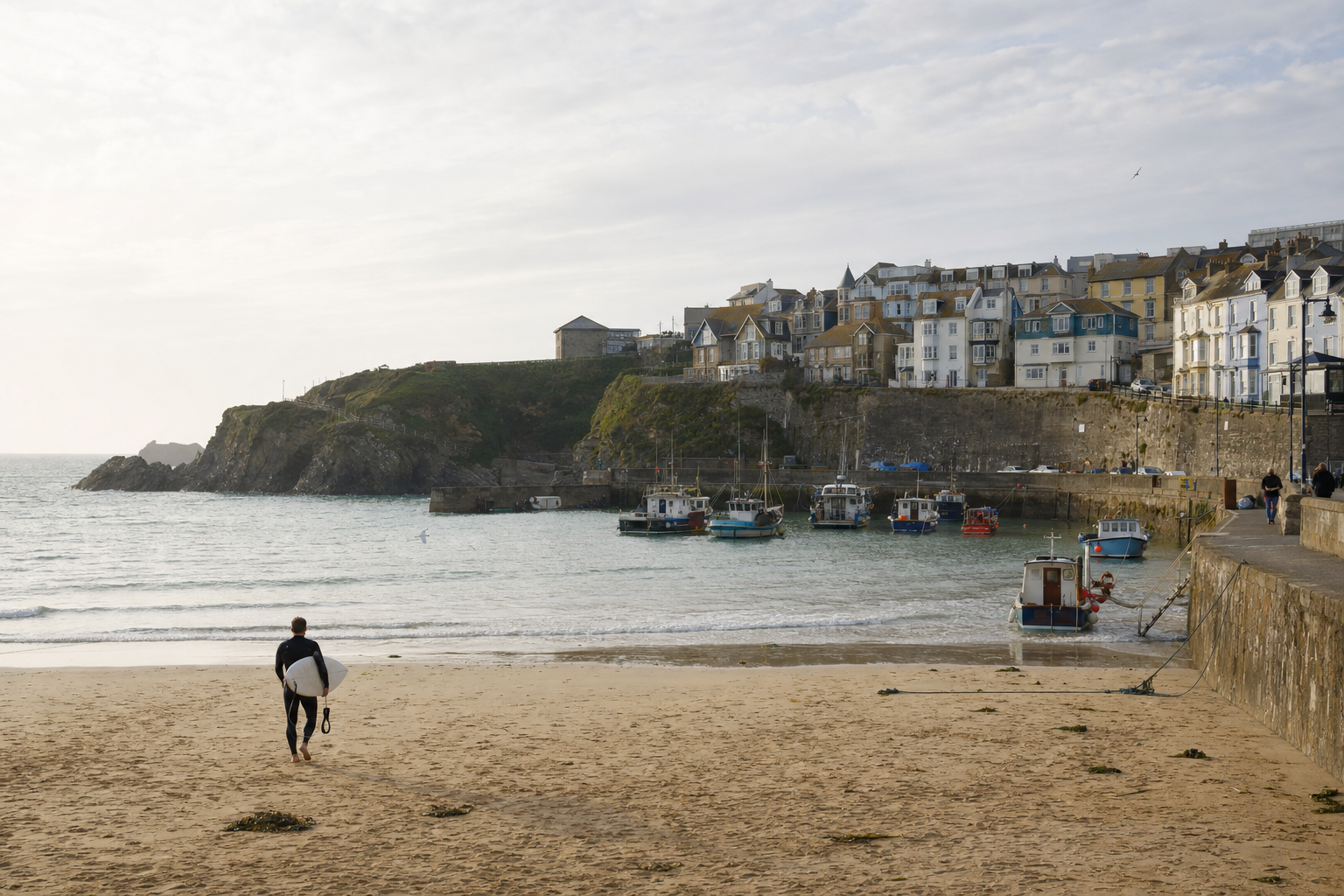 A surfer walks along a sandy beach towards the sea, with boats docked and a coastal village in the background.