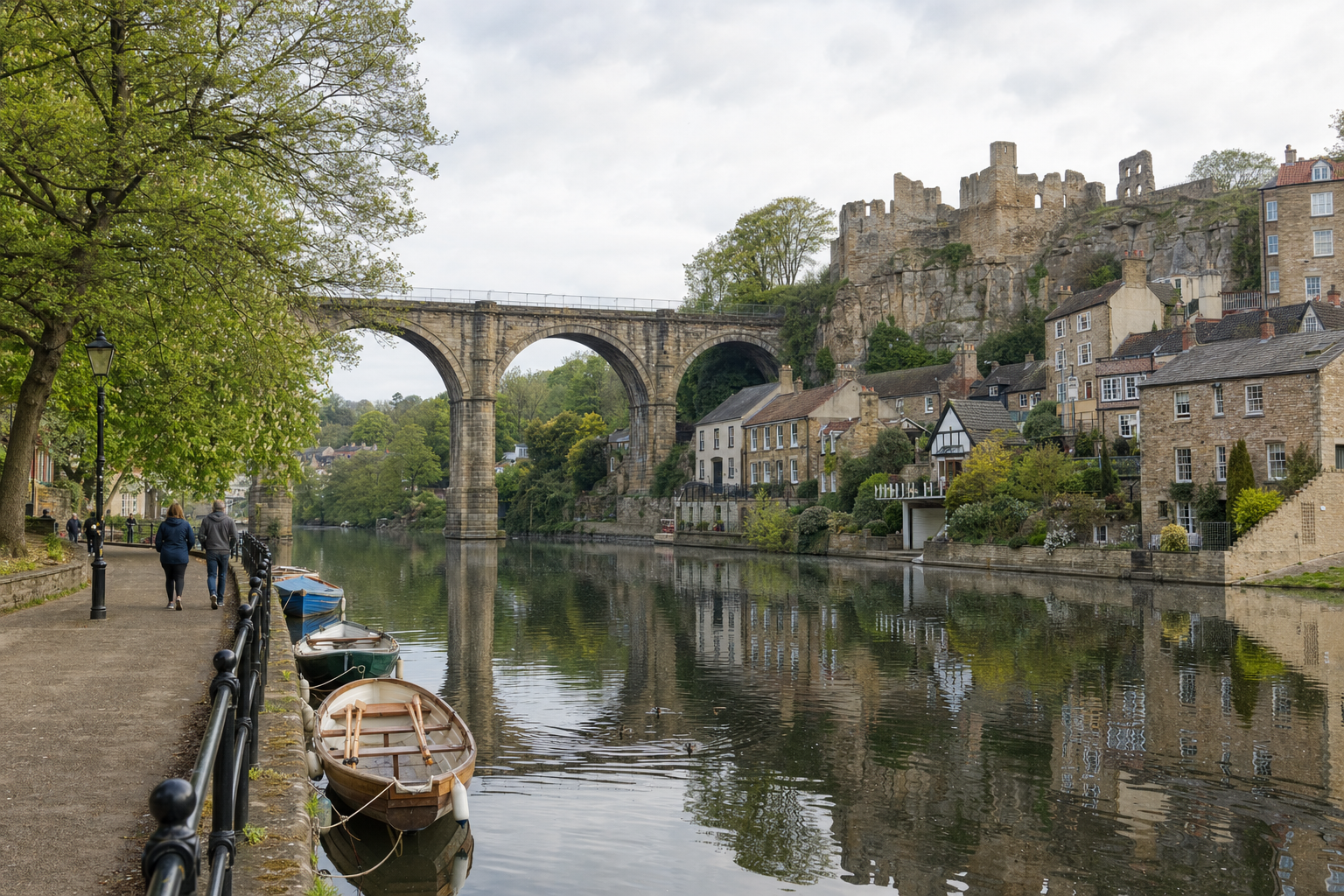 A riverside path with people walking, boats moored, and a stone viaduct with castle ruins above.