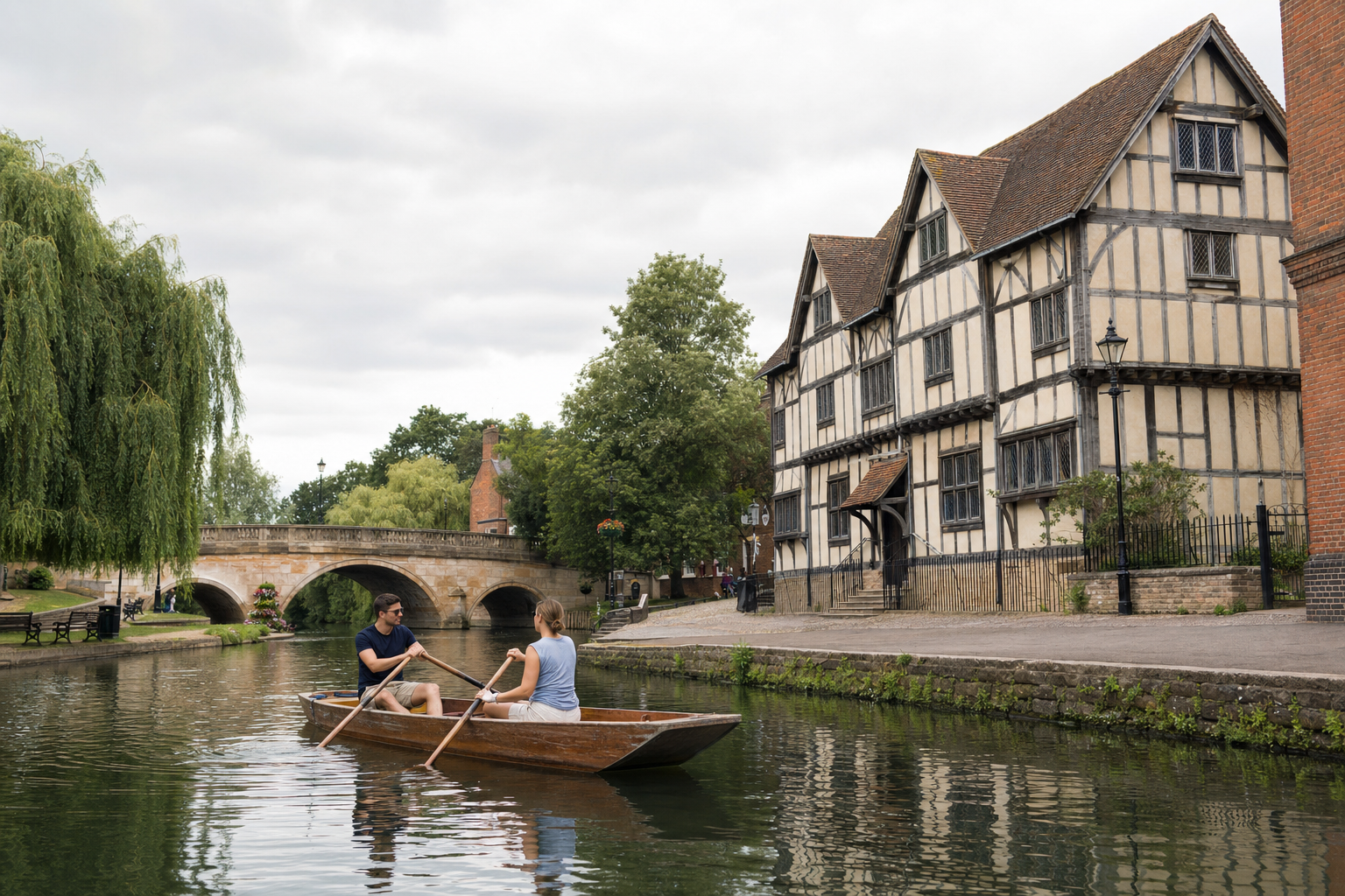 Two people rowing a wooden boat on a tranquil river beside a historic half-timbered house and arched stone bridge.