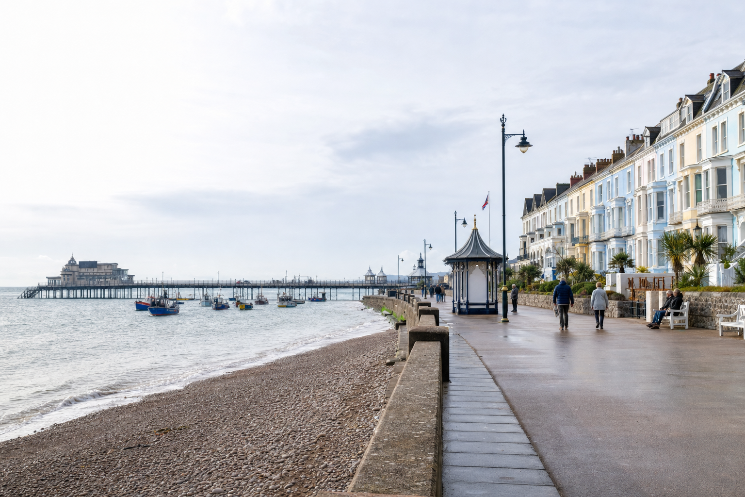 Seaside promenade with colourful Victorian houses, a pebble beach, and a pier extending into the sea under a cloudy sky.