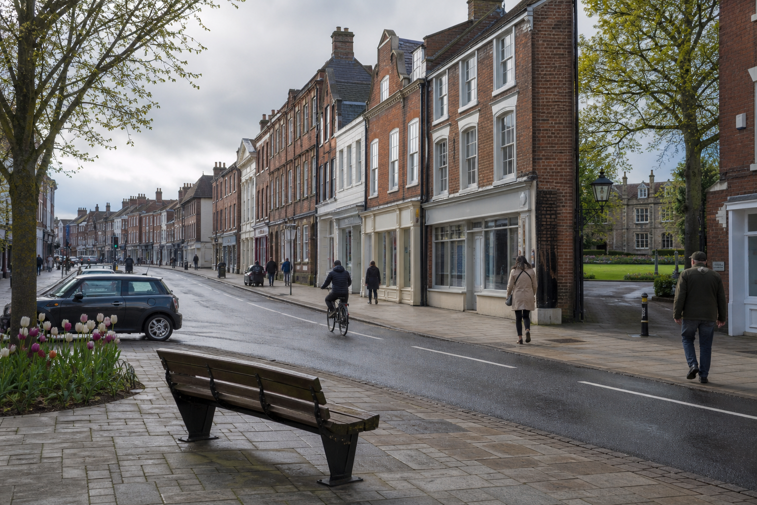 A cyclist rides down a wet street lined with historic terraced buildings, with pedestrians walking by and tulips in bloom.