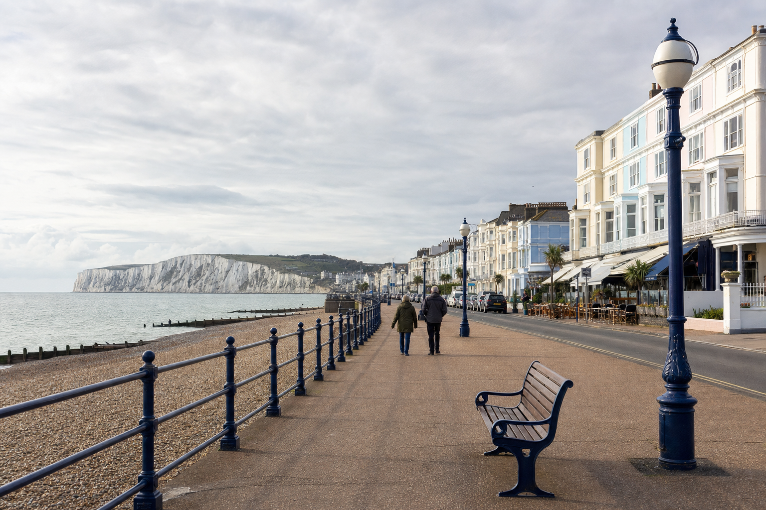 Two people walk along a seaside promenade with cliffs and Victorian buildings in the background.