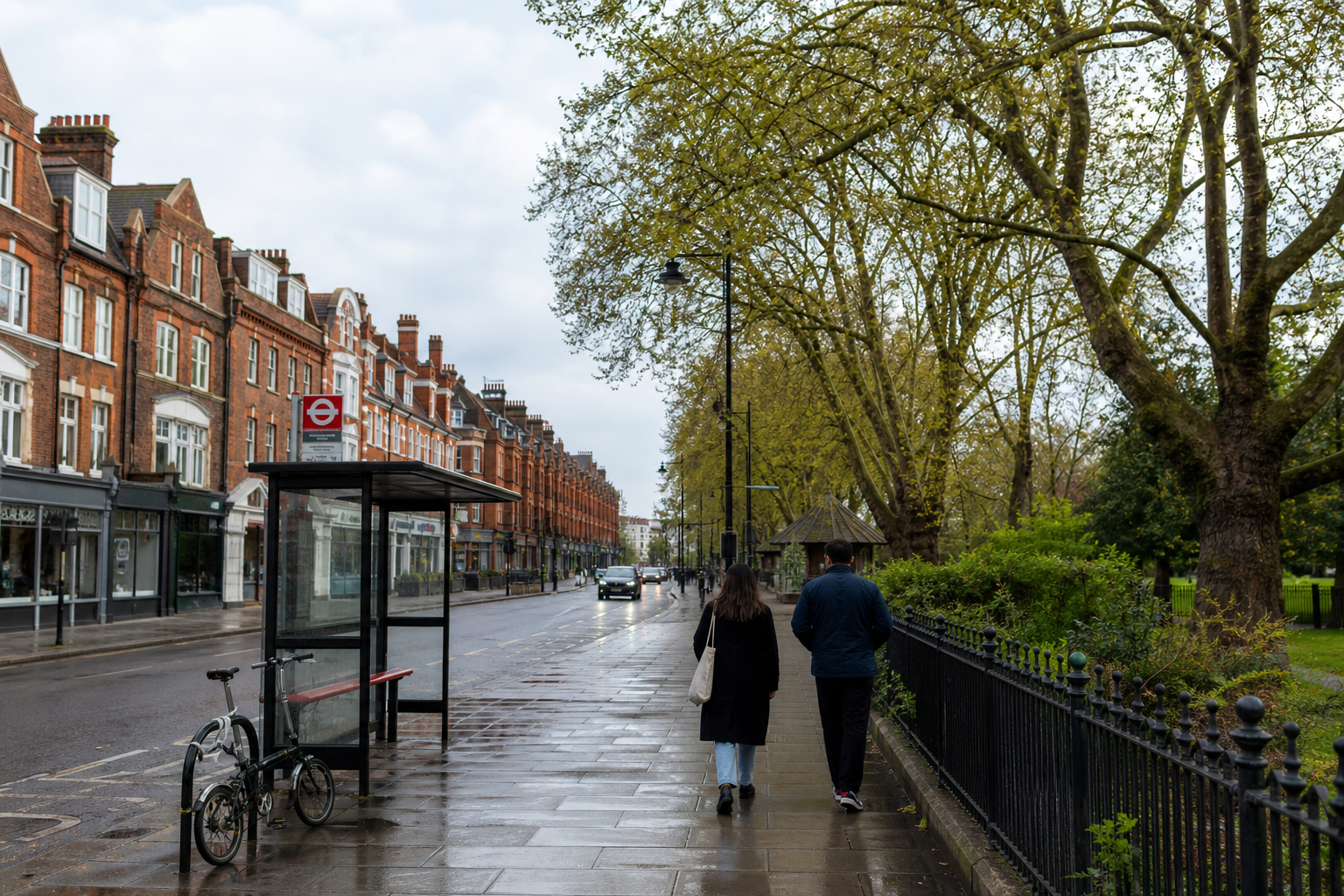 A couple walks along a wet sidewalk past a bicycle and bus stop on a tree-lined street.