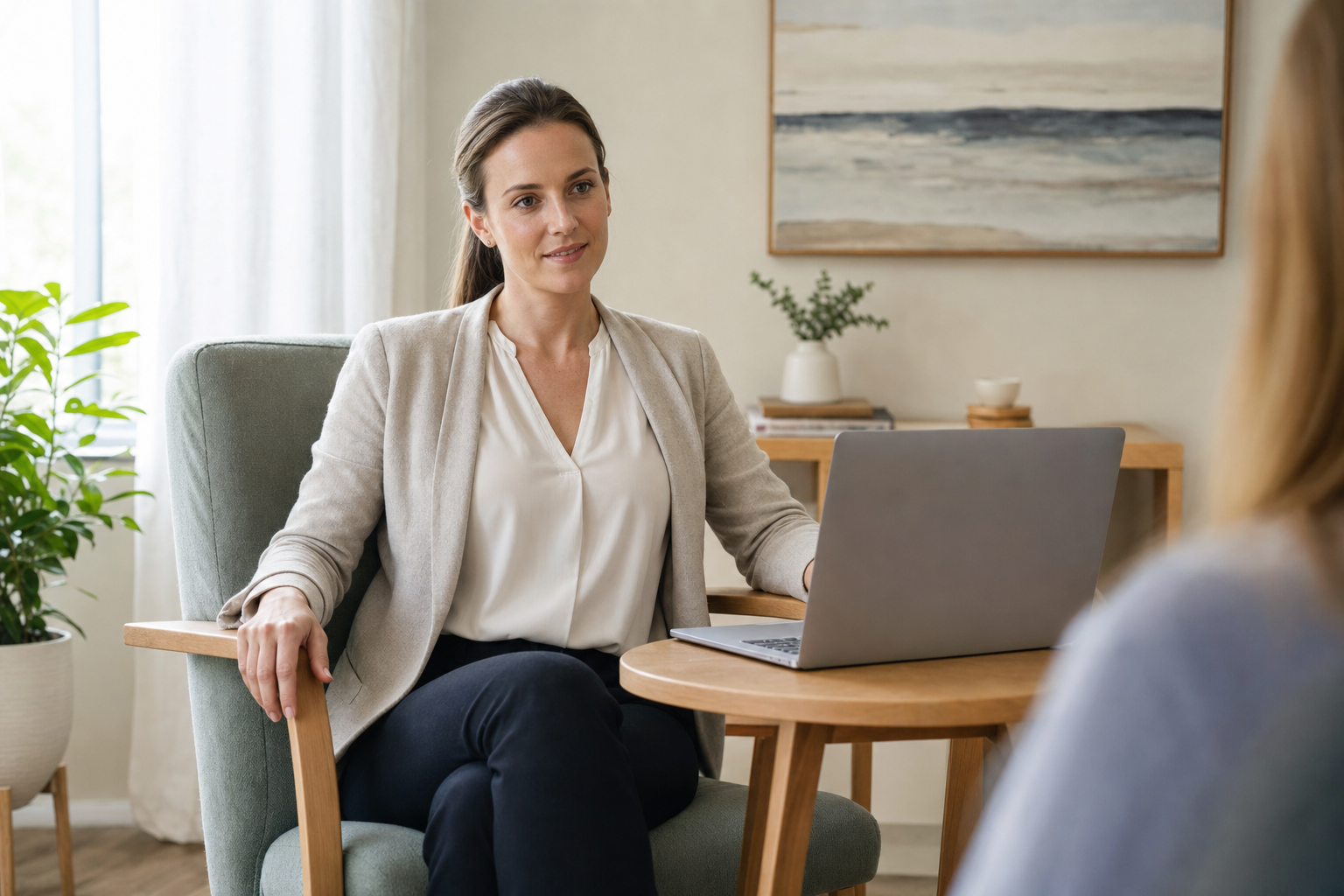 A woman in a blazer sits in an armchair, listening attentively with a laptop open on the table beside her.