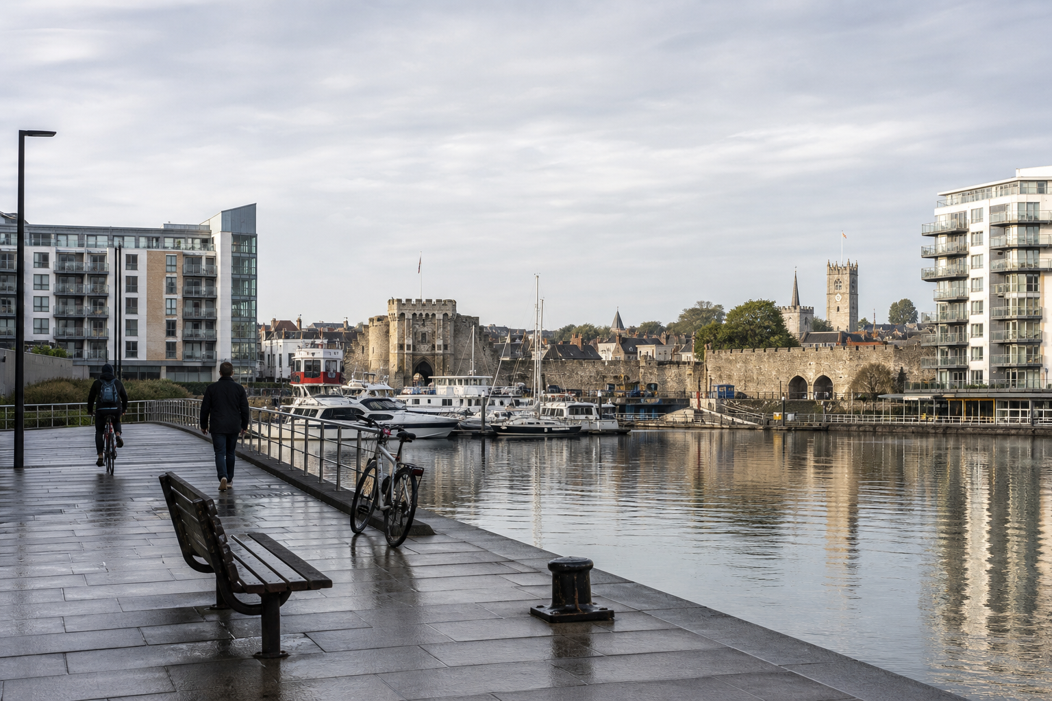 Waterfront walkway with people and bicycles alongside a marina, historic buildings, and modern apartments.