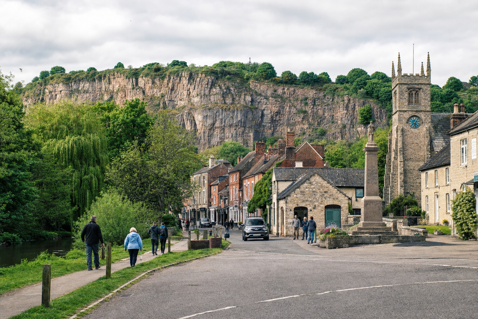 A scenic village street with a stone church, war memorial, and people walking against a backdrop of a cliff and trees.
