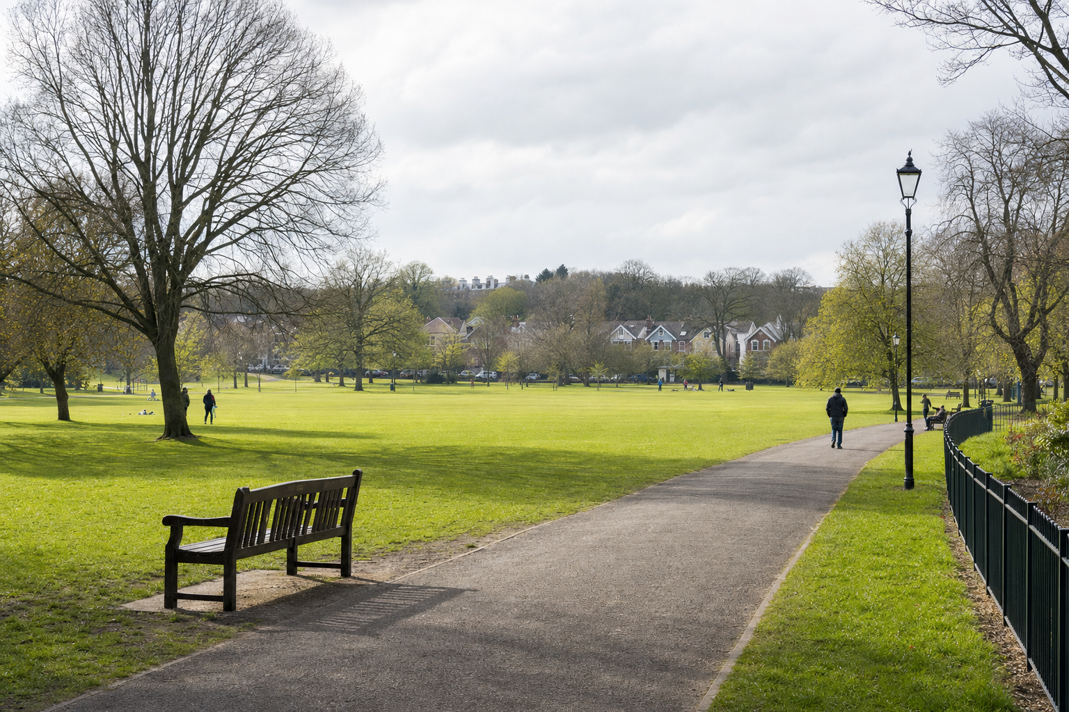 A peaceful park scene with a path, benches, people walking, and houses in the distance on a cloudy day.