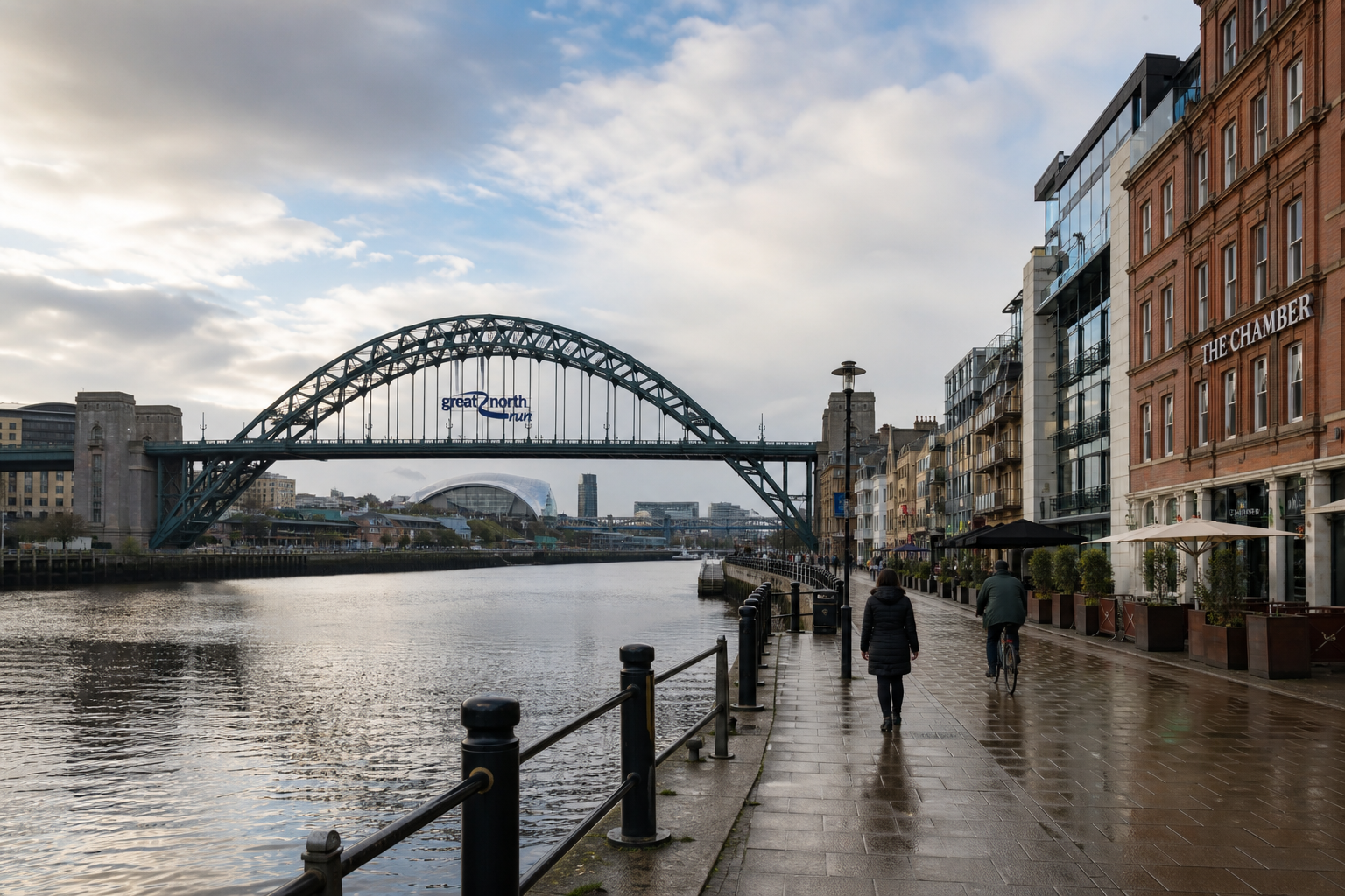 People walk along a riverside path with a large arched bridge and cityscape in the background.