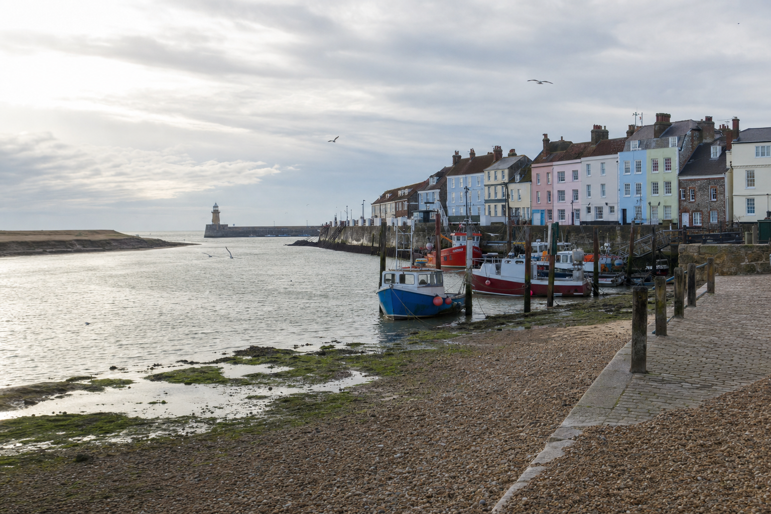 Fishing boats moored in a harbour next to colourful terraced houses with a lighthouse in the distance.