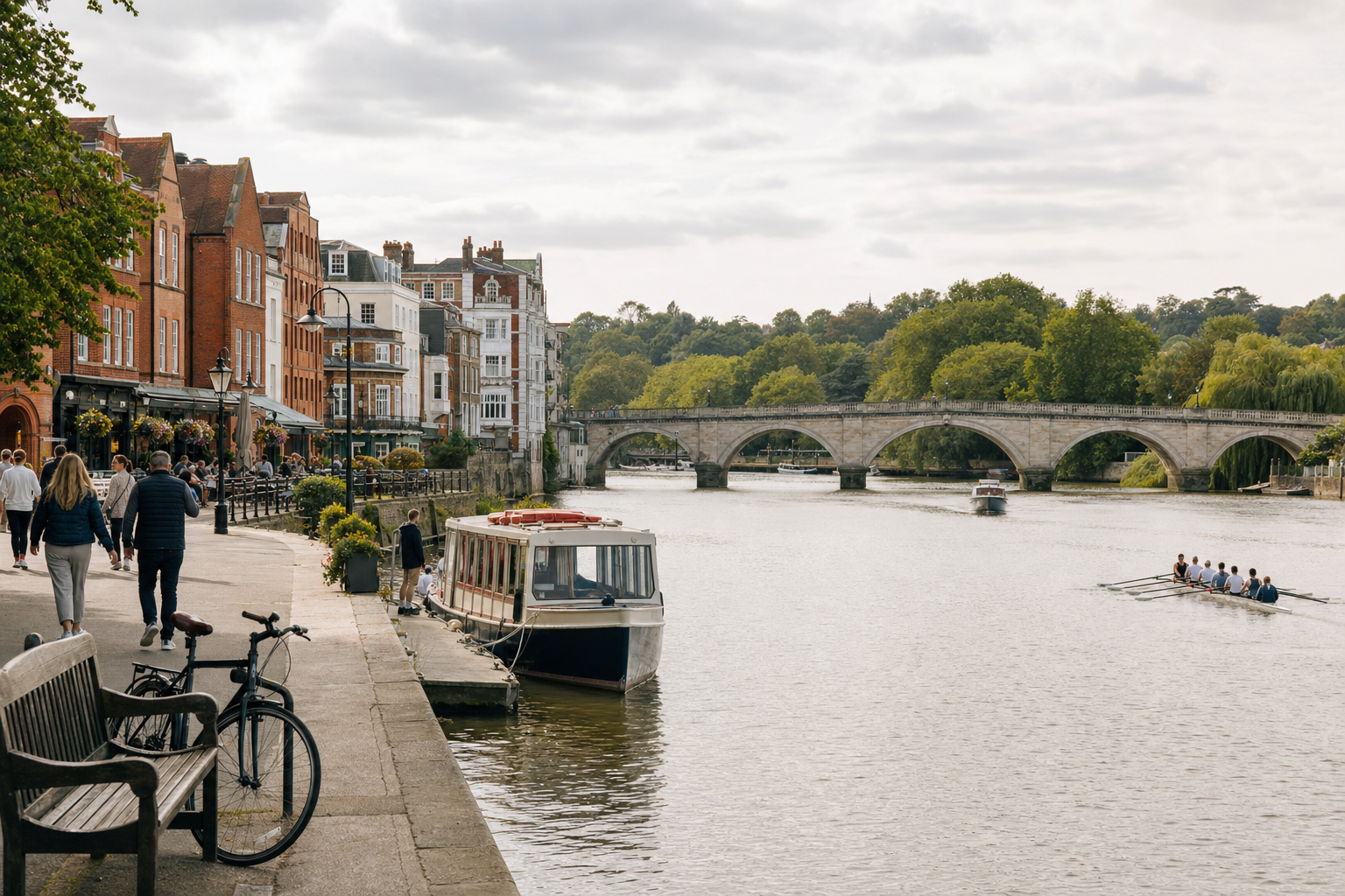 People stroll along a riverside promenade with boats docked and rowers on the river, near an arched stone bridge.