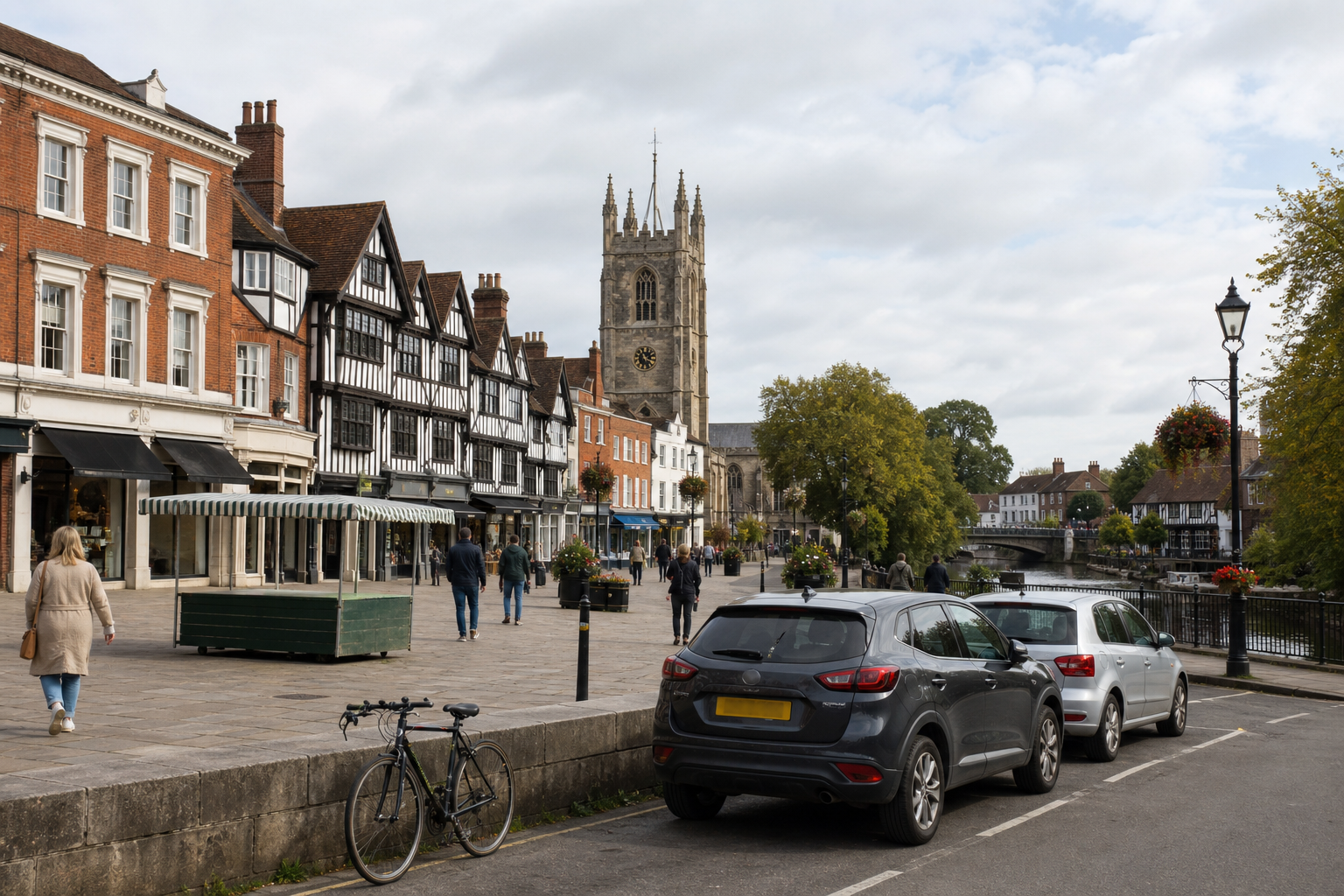 Historic English street with half-timbered buildings, a church tower, parked cars, and people walking along the riverbank.