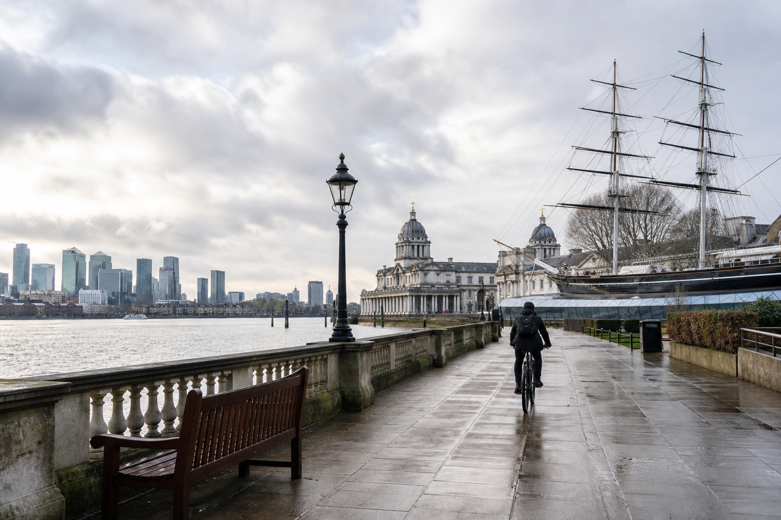 Cyclist rides along Thames riverside with historic buildings and a ship in the background under a cloudy sky.