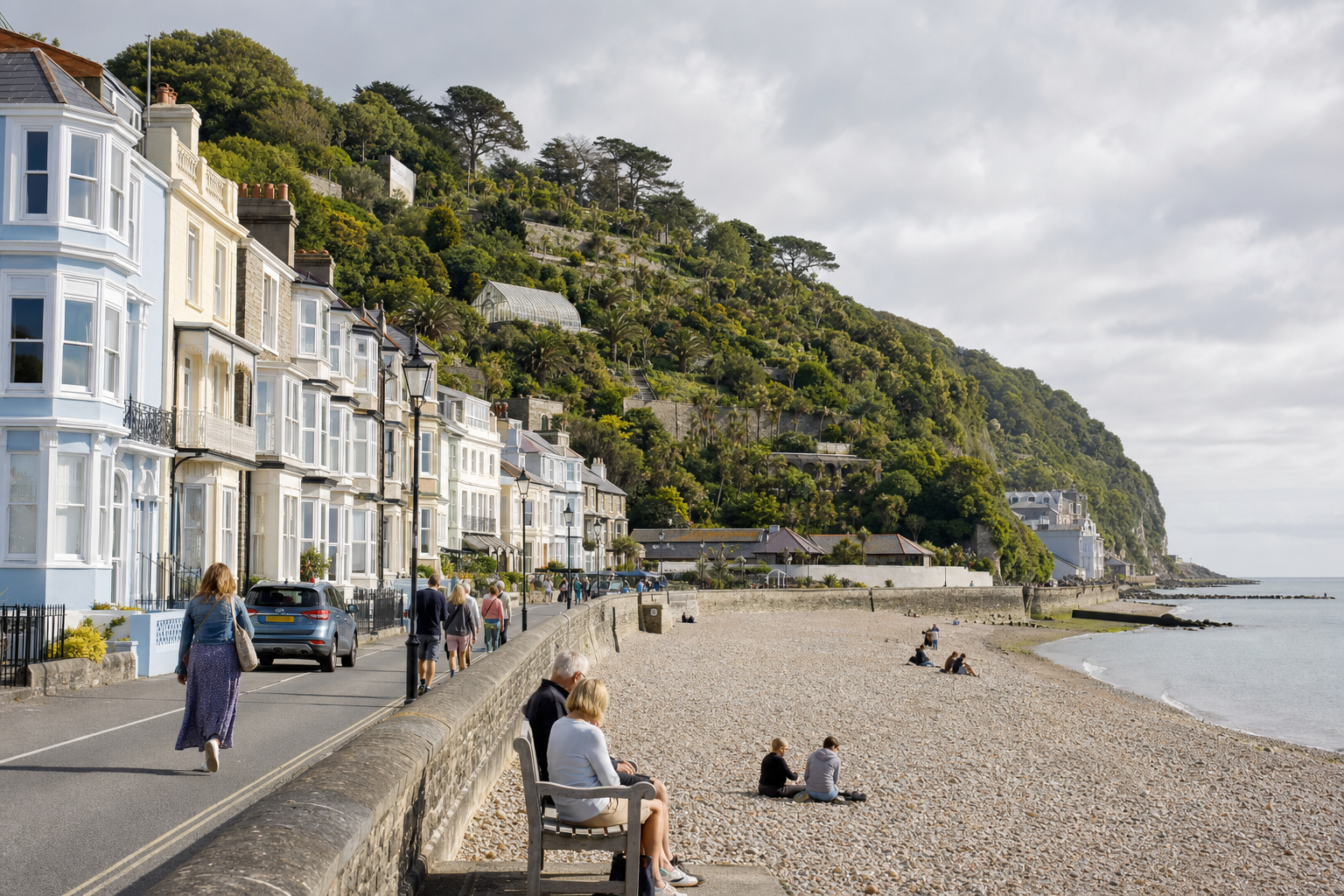 People walking along a promenade beside a pebbled beach with a row of Victorian houses and lush green hillside.
