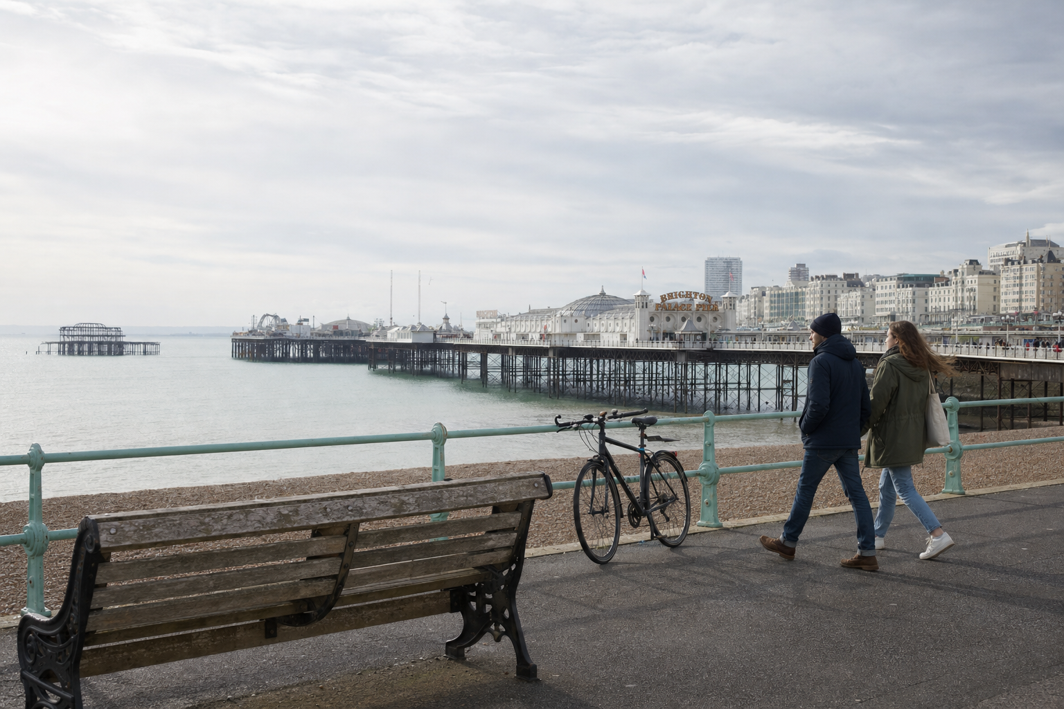 Two people walking along Brighton seafront with a bike propped against the railing and the pier in the background.