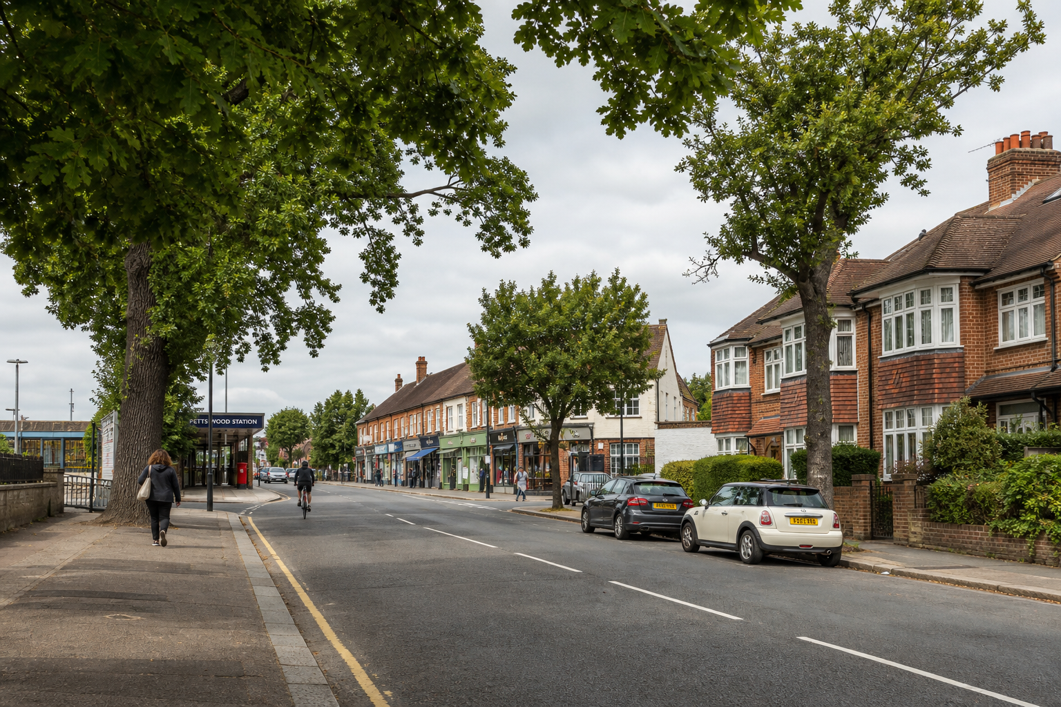 Tree-lined street with shops on one side, houses on the other, and a few pedestrians near a railway station entrance.