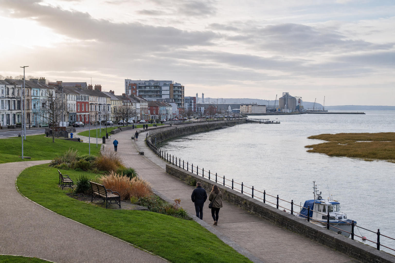 People walking along a promenade by the water in a coastal town, with colourful houses and a dock visible.