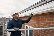 A construction worker in a hard hat inspects the roofline of a brick building while standing on scaffolding.