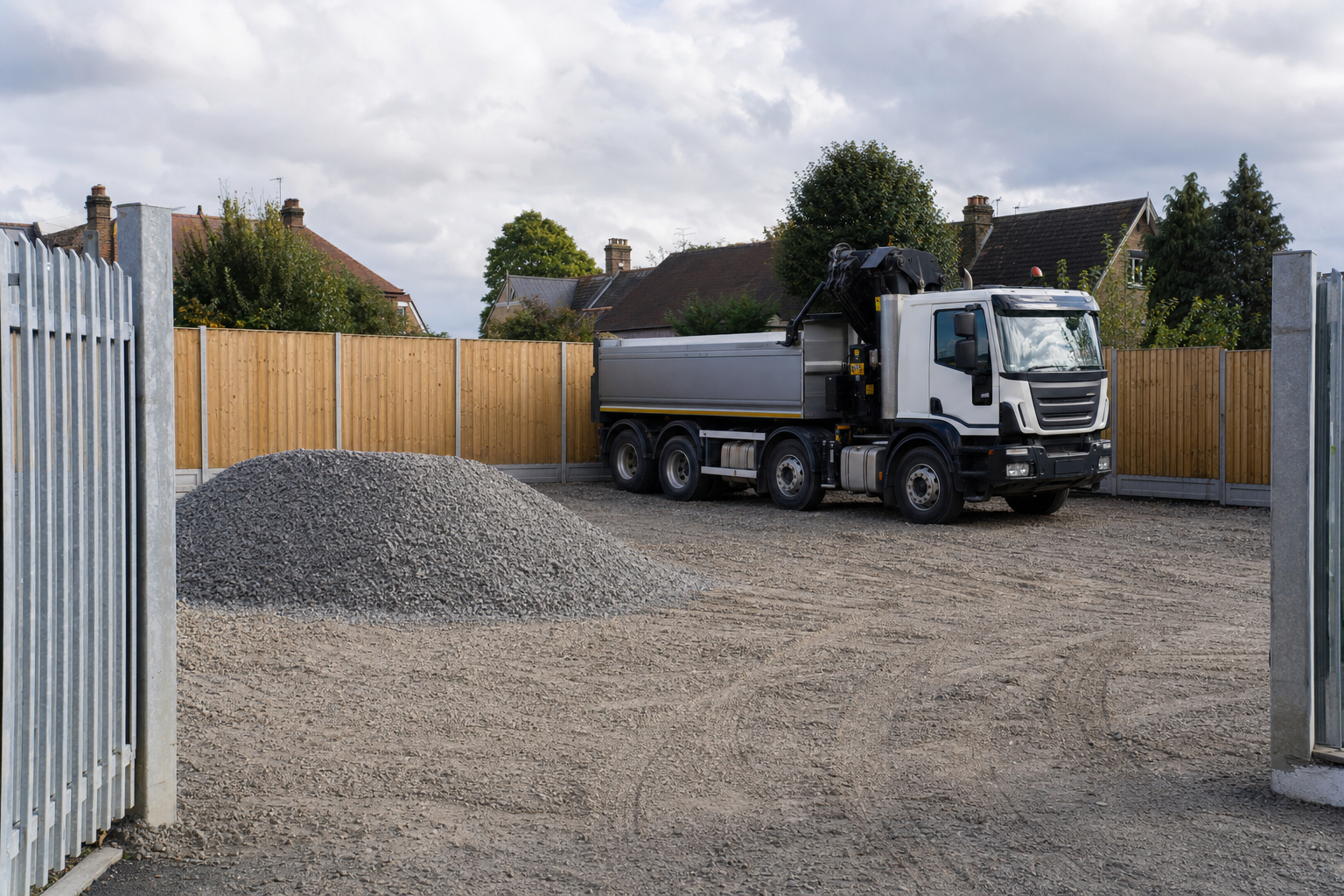 Lorry parked on a gravel lot next to a wooden fence in a residential area with houses and trees in the background.