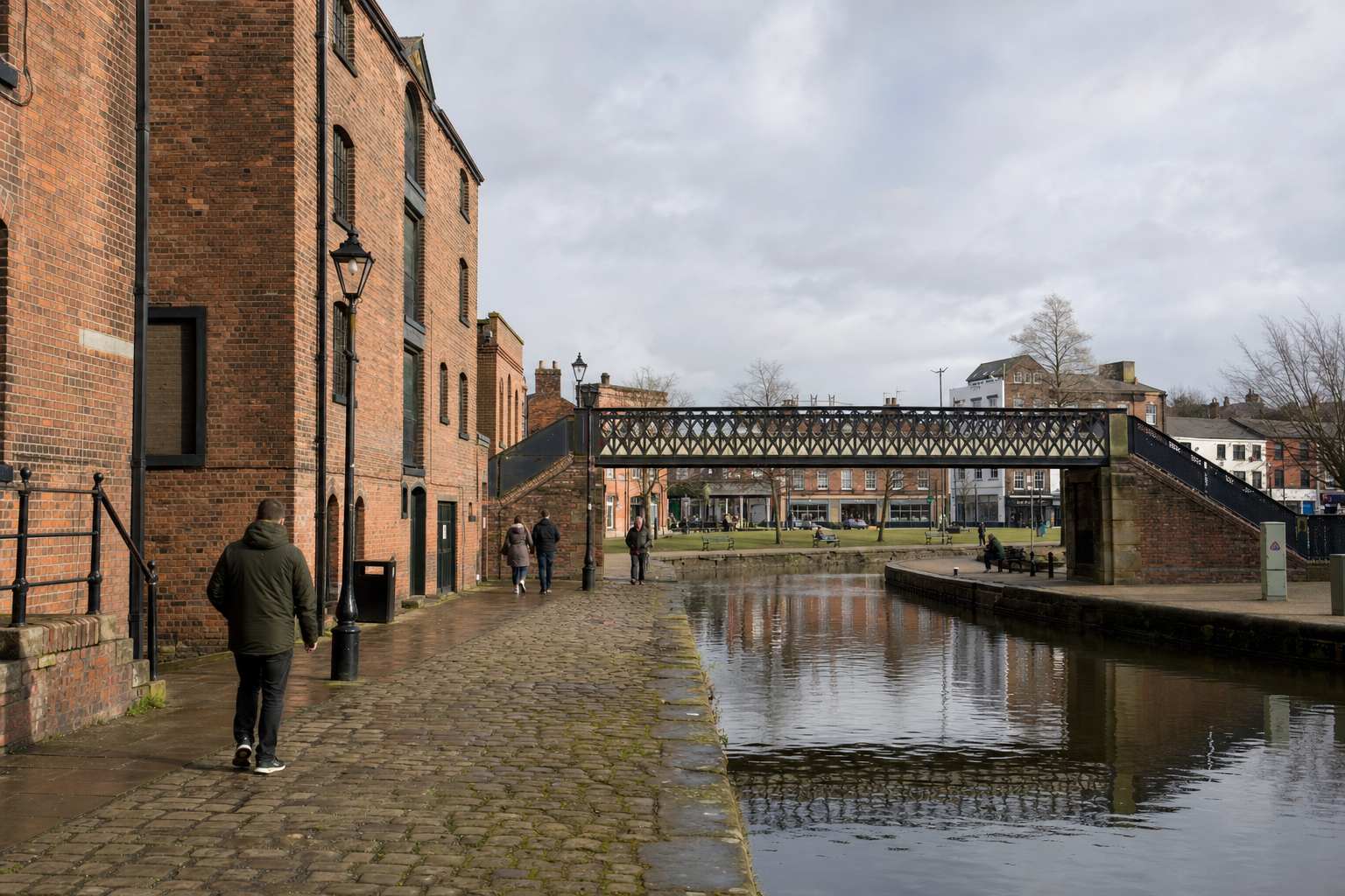 A cobblestone path runs alongside a canal beneath a Victorian-style iron bridge, with people walking nearby.