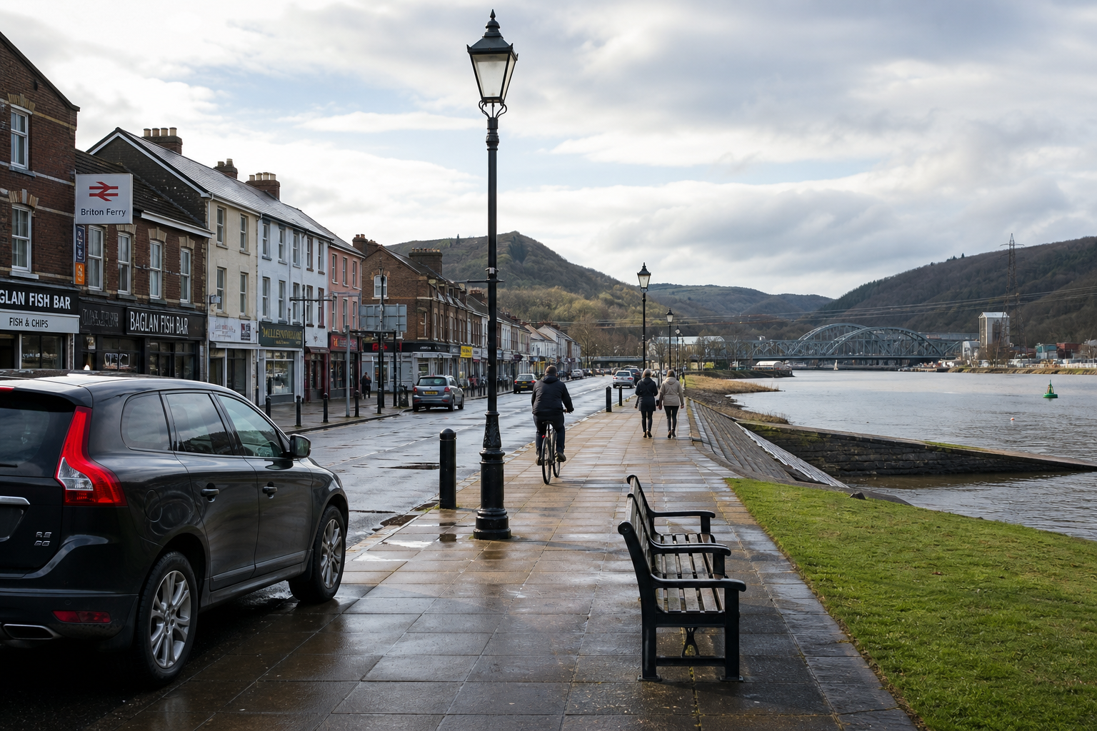 People walking and cycling along a riverside promenade with shops, cars, and distant hills.