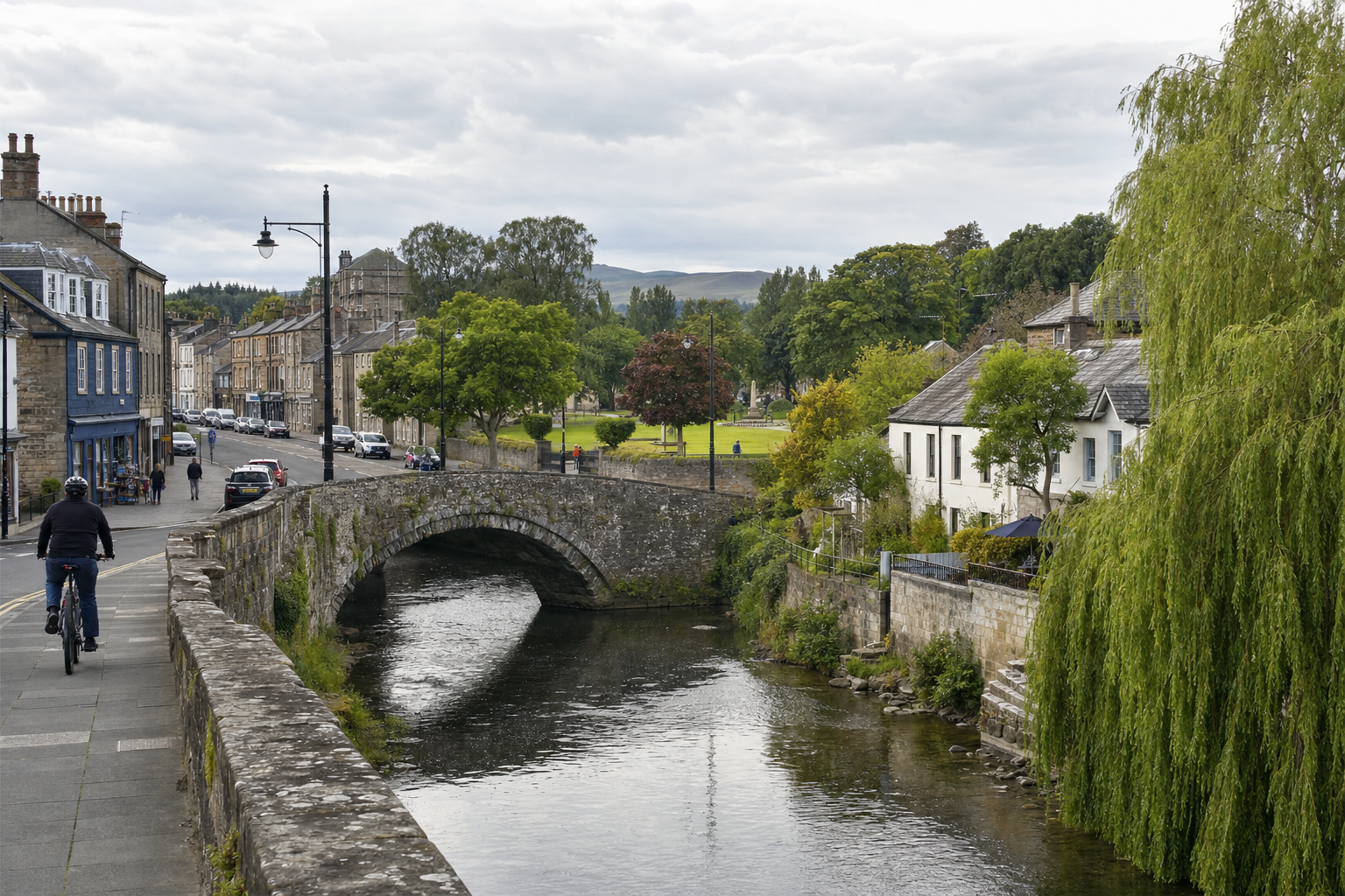 A cyclist rides along a riverside path beside a stone bridge in a picturesque village with green trees and historic buildings.
