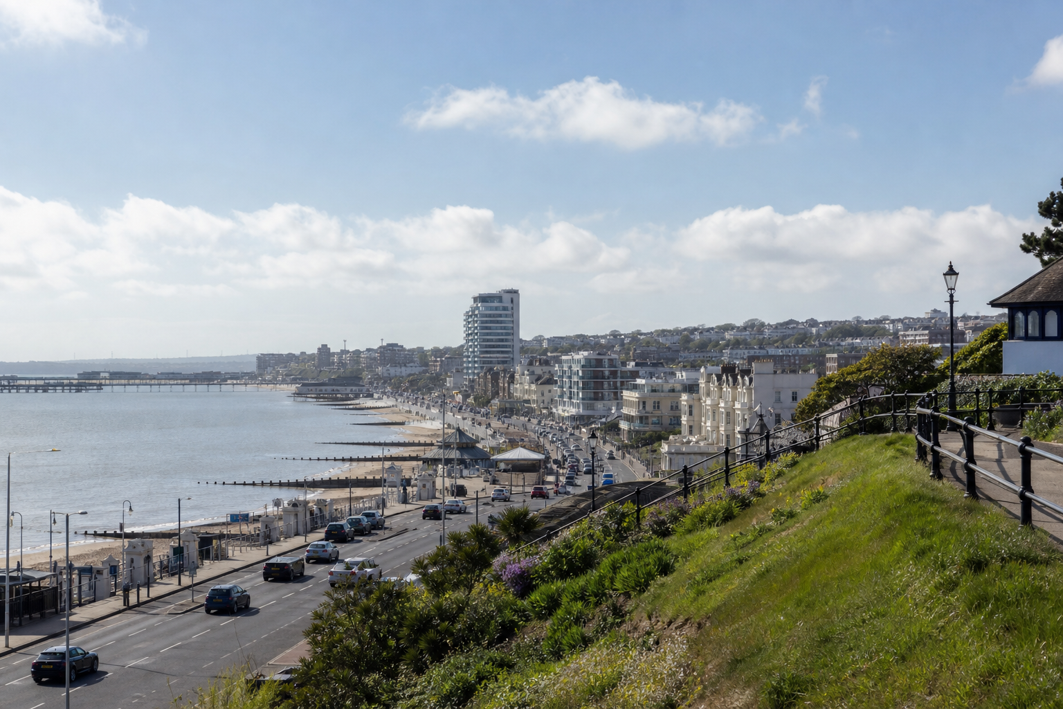 Seaside road lined with cars and buildings on a sunny day, with a grassy hill in the foreground.