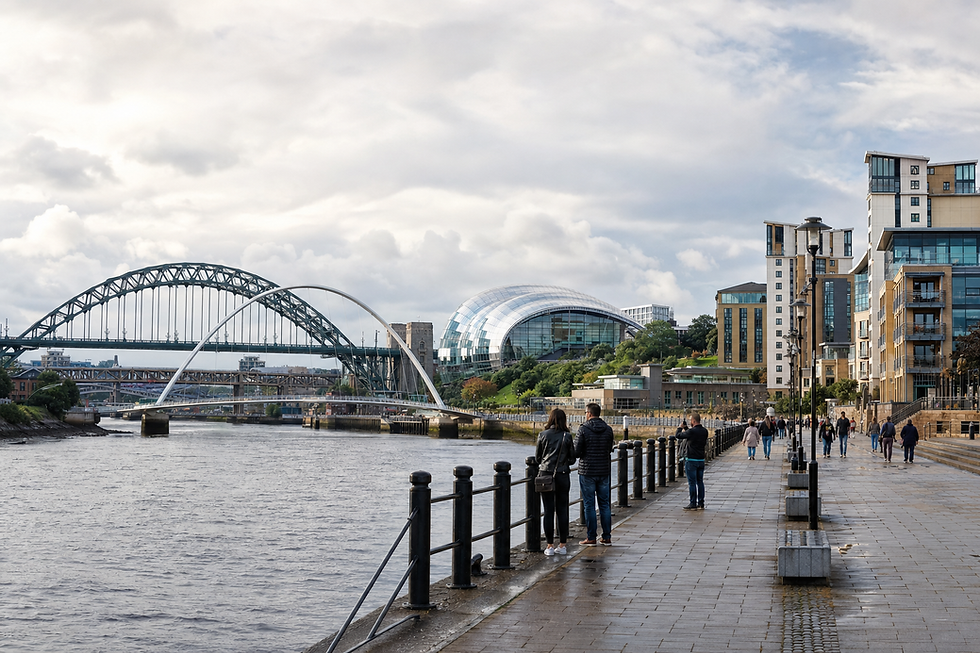 People walking along a riverside promenade with bridges and a modern building in the background under a cloudy sky.