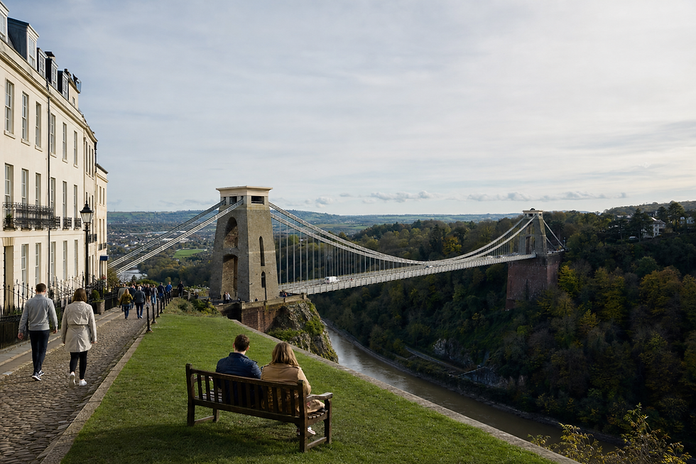 Clifton Suspension Bridge spans across a scenic gorge with people walking nearby and a couple seated on a bench.