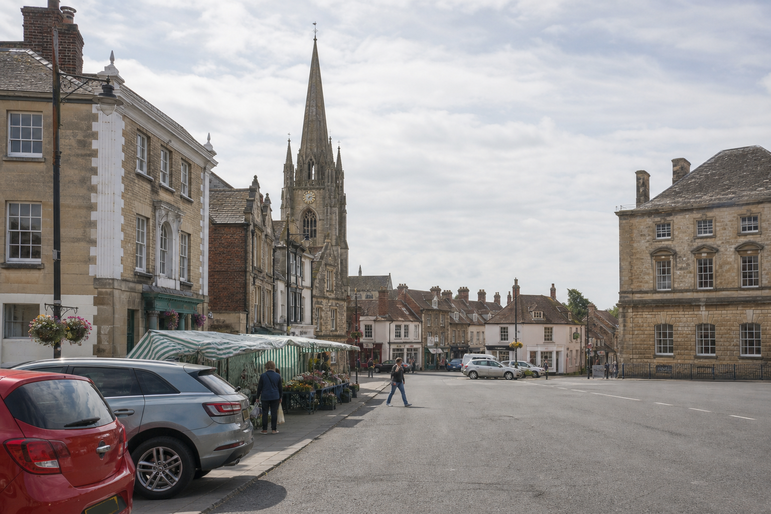 A historic town square with a church spire, market stalls, and people walking near parked cars under a cloudy sky.