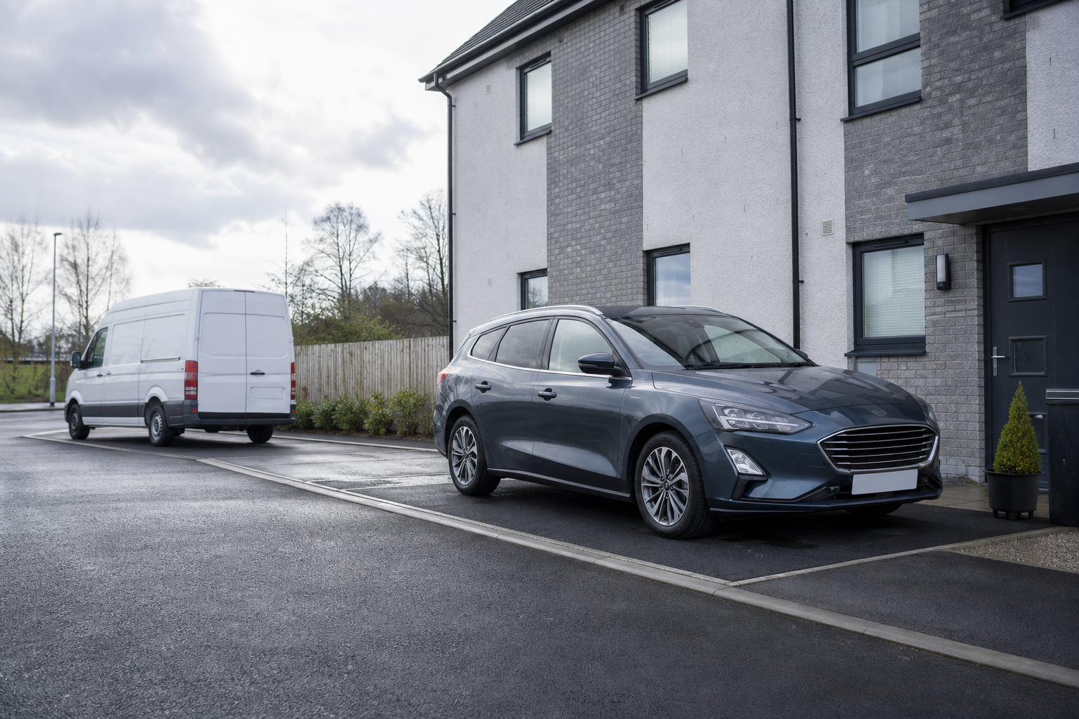 A grey estate car is parked in a driveway next to a modern house, with a white van on the street nearby.