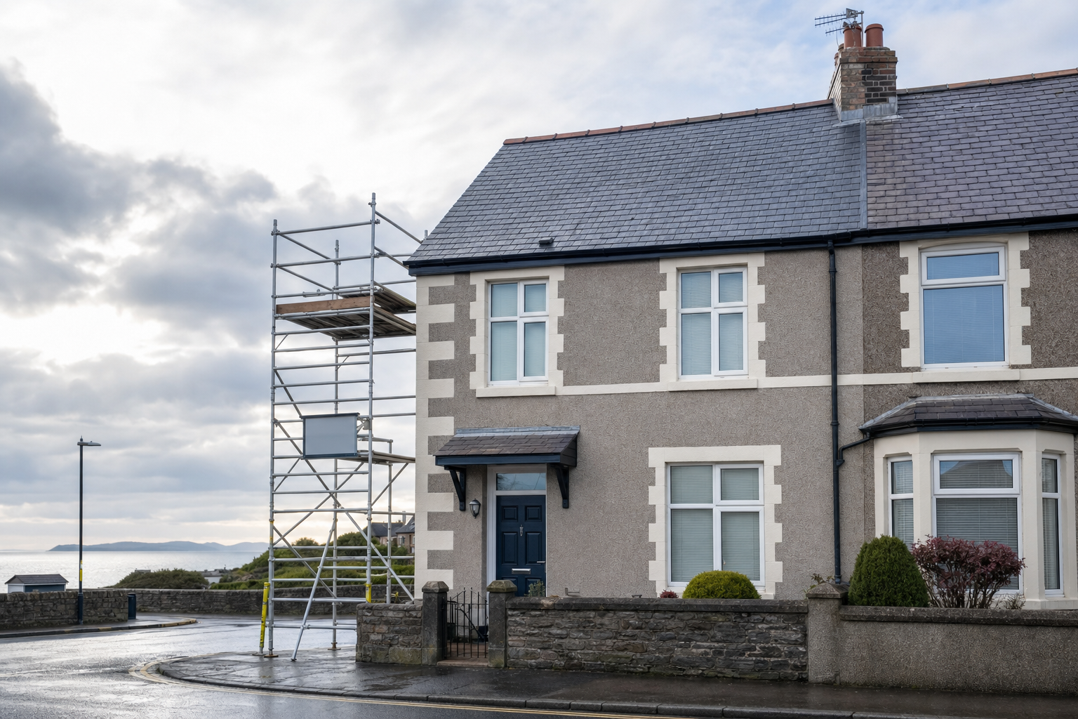 A scaffolding setup stands next to a semi-detached house near a coastal road under a cloudy sky.