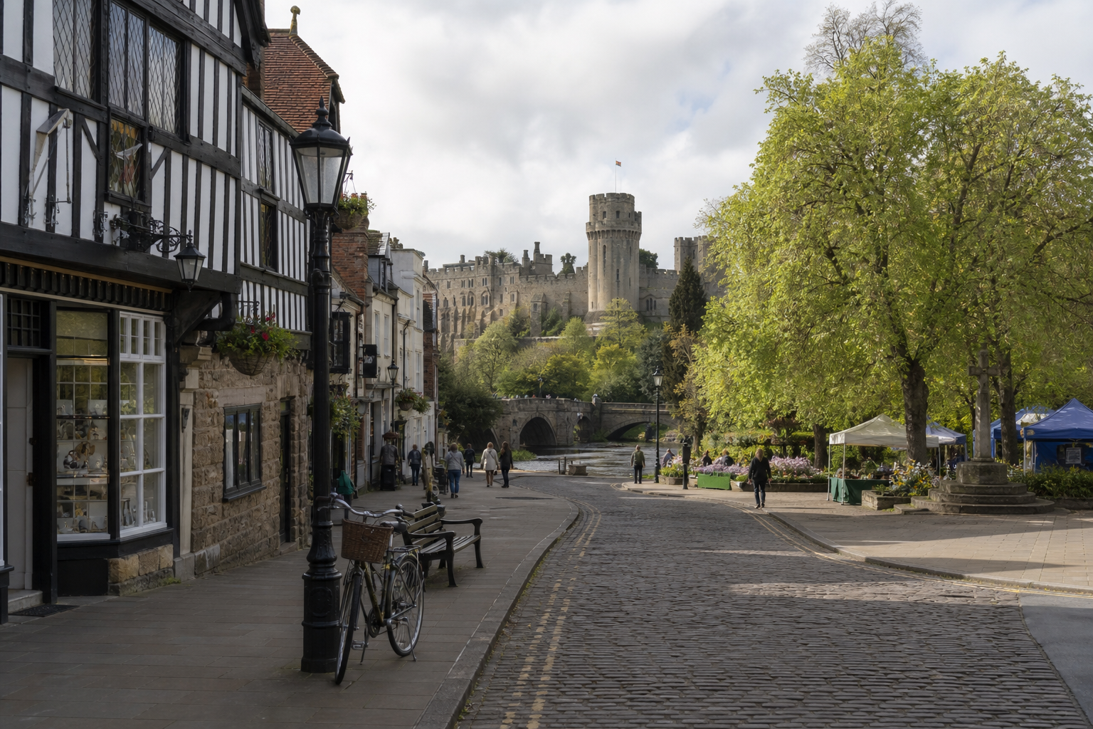 Cobblestone street in a historic town with a view of a medieval castle, timbered houses, and lush trees under cloudy skies.