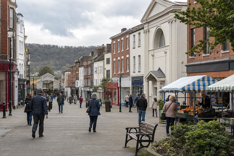 Pedestrians walk along a bustling high street beside market stalls in front of historic buildings on a cloudy day.