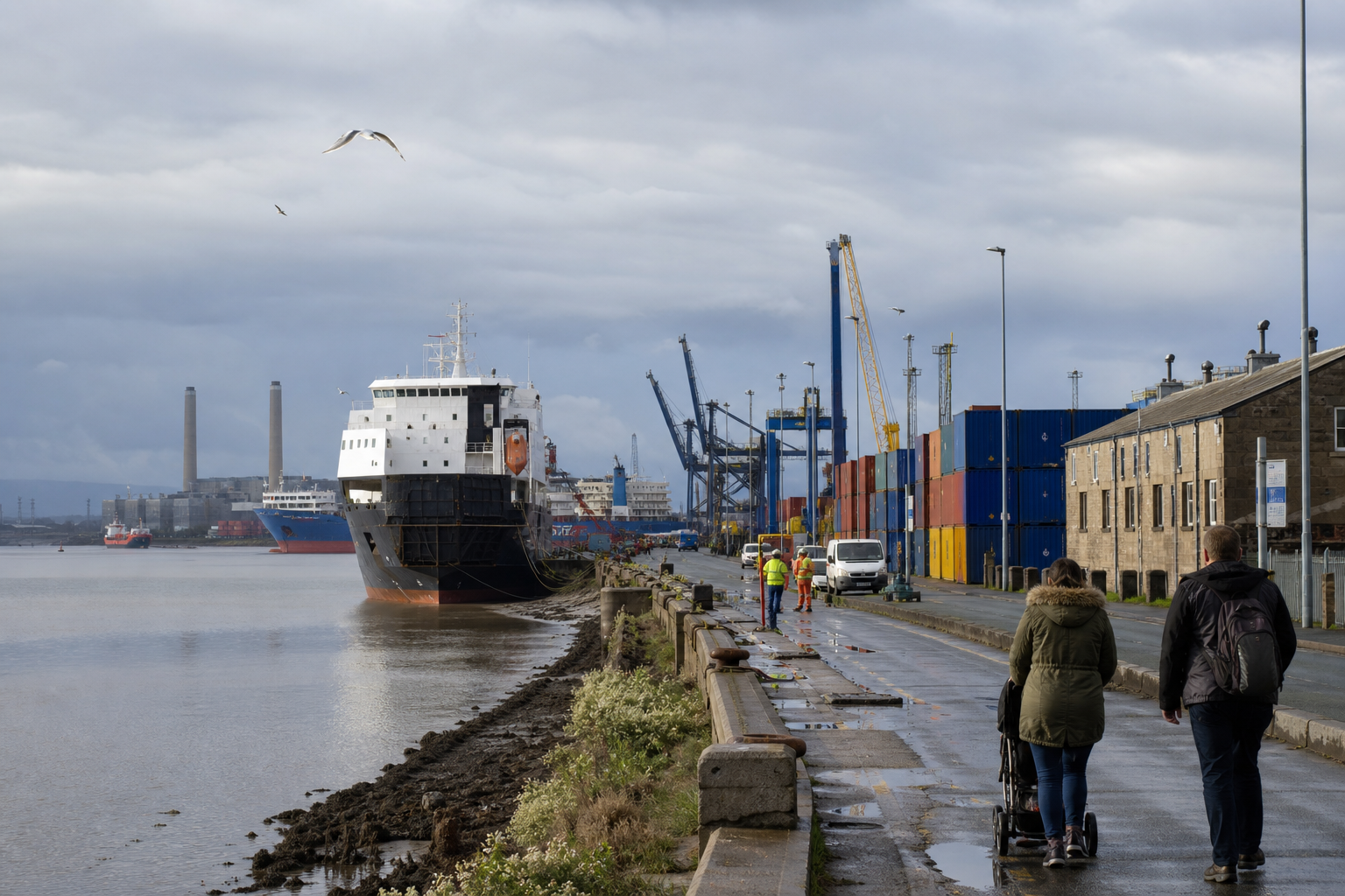 Cargo ship docked at an industrial port with people walking along the waterfront.