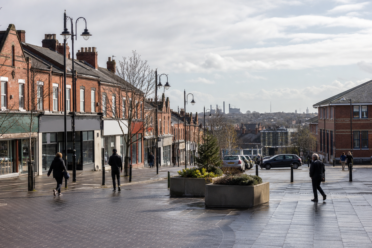 People walking on a sloped street lined with red brick buildings and leafless trees under a cloudy sky.