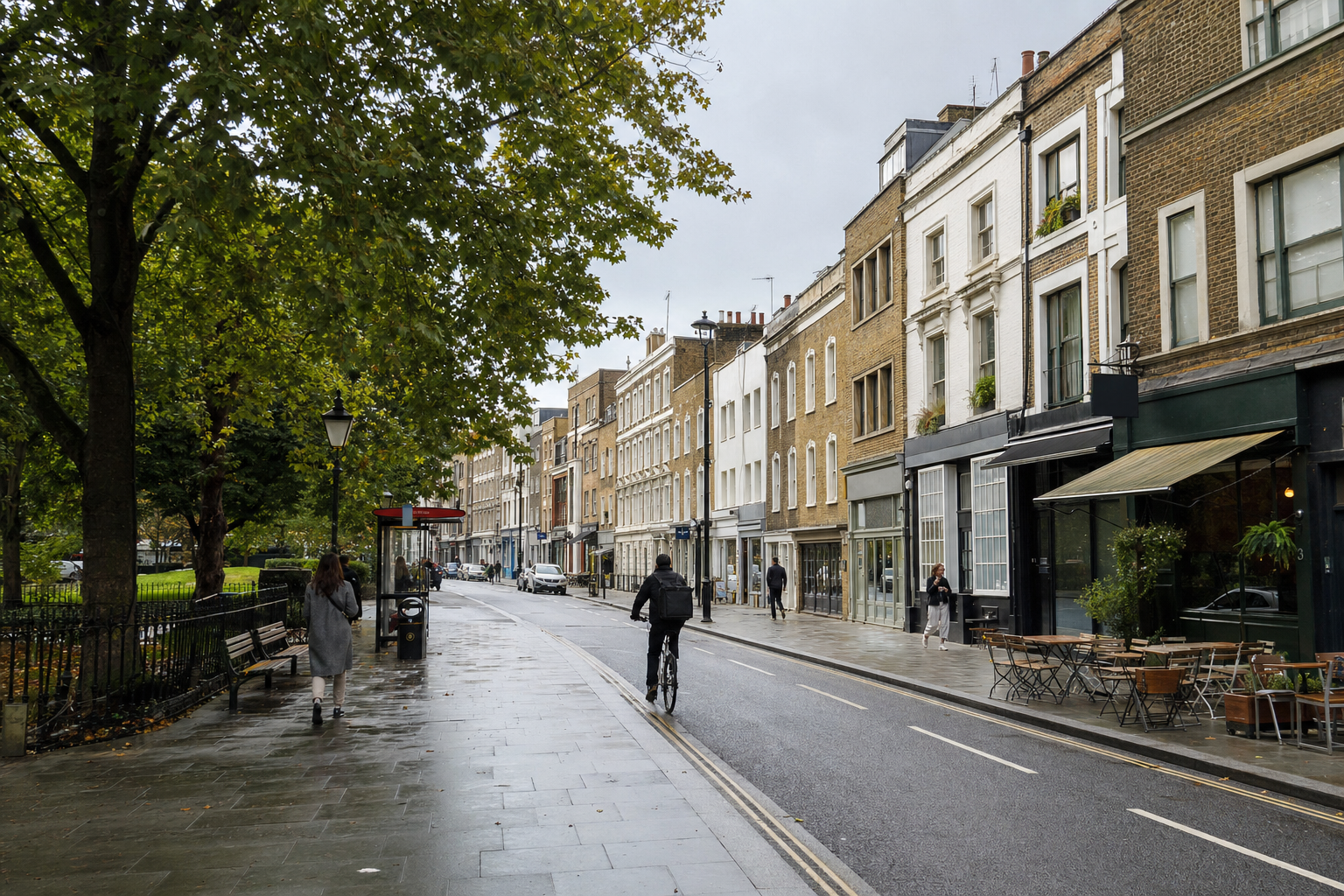 A cyclist rides along a wet street lined with trees and terraced buildings on a cloudy day.