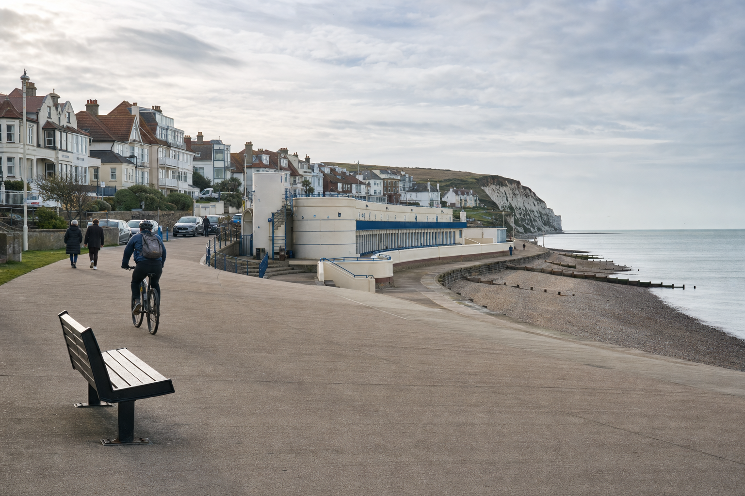 Cyclist riding along a quiet seafront promenade next to pebble beach and historic buildings.