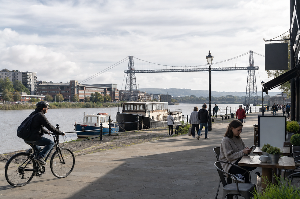 A cyclist rides along a riverside path with pedestrians and a seated woman using her phone, near a bridge in the background.
