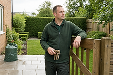 Man in a green fleece holding gloves, leaning on a wooden gate in a neatly landscaped garden.