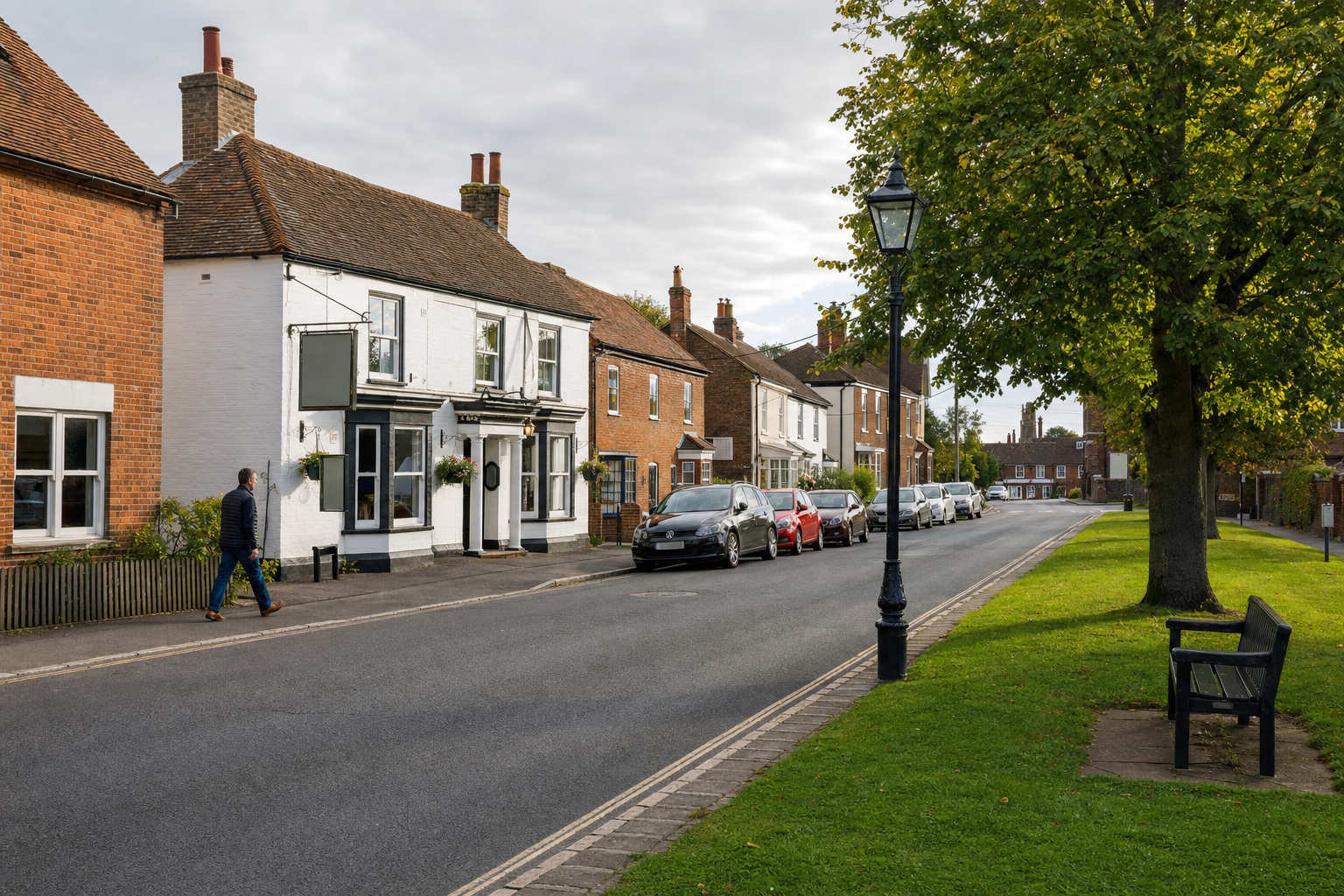 A quiet street scene with a man walking past traditional buildings and parked cars, lined with a green verge and trees.