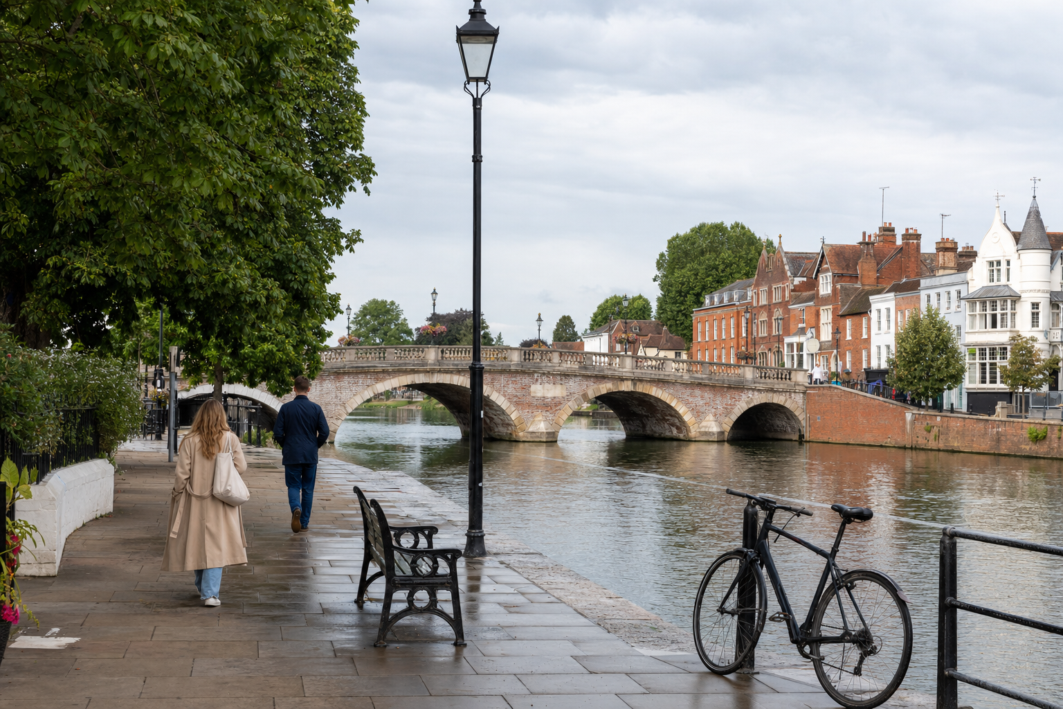 A riverside path with people walking, a bicycle, and a historic stone bridge in the background.