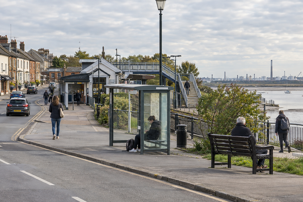 A quiet riverside street features a bus shelter, pedestrians, benches, and distant industrial structures across the water.