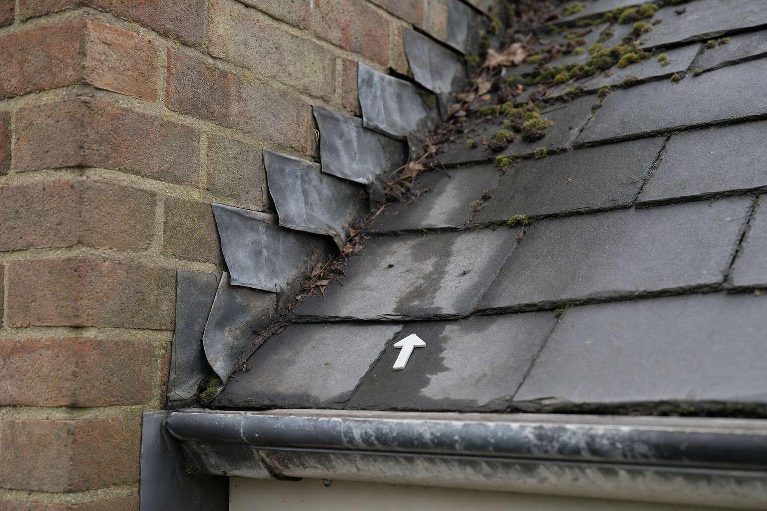 Moss and debris accumulate in the gap between a brick chimney and slate roof with an arrow marking a specific tile.