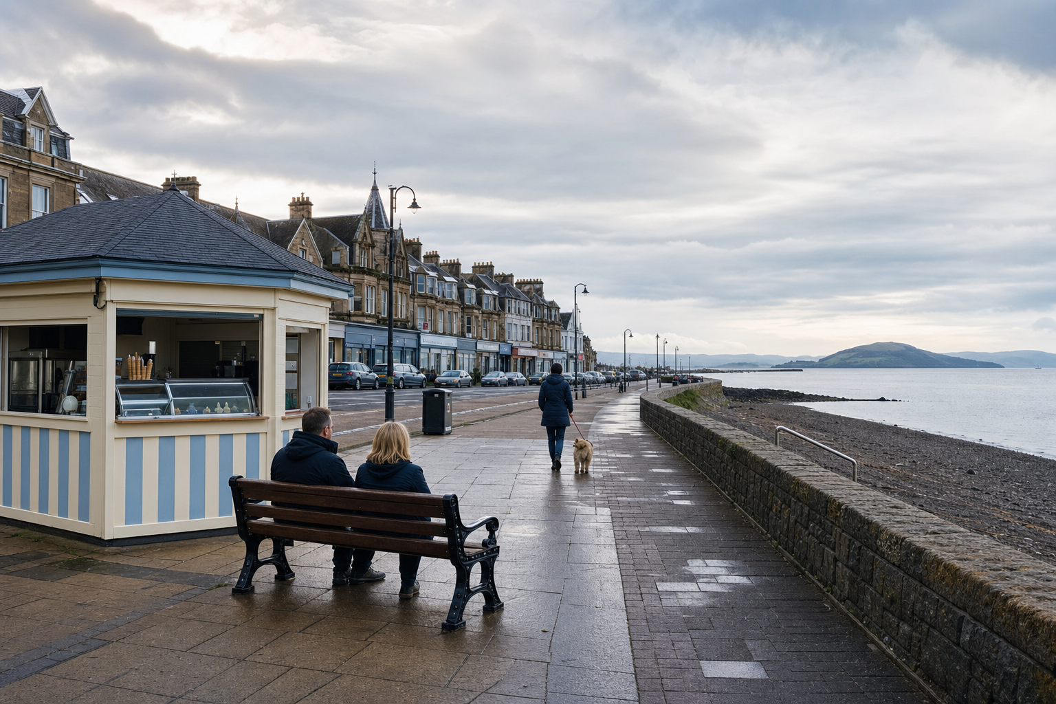 People sitting on a bench near a beachside kiosk, with a promenade and distant hills under a cloudy sky.