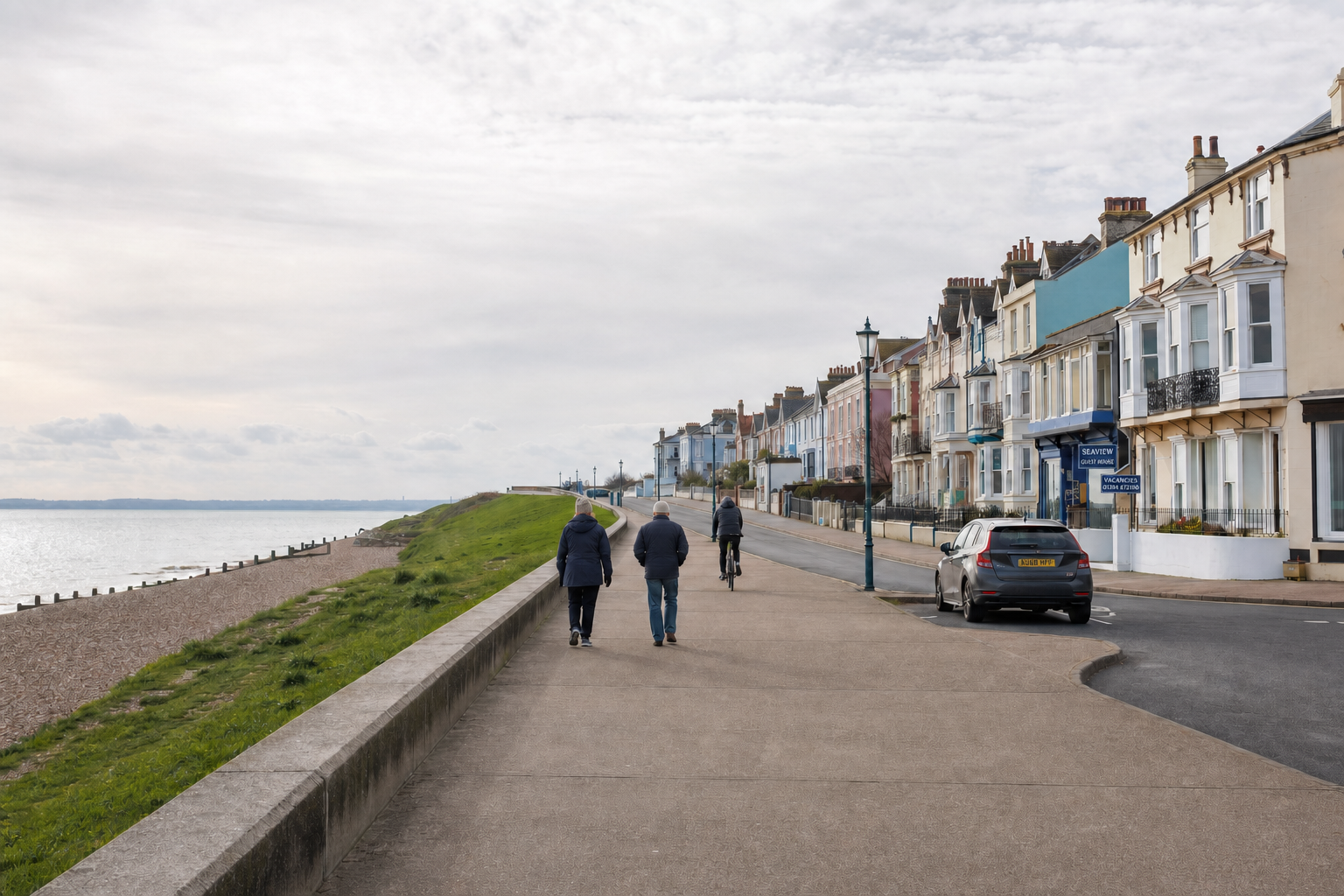 People walking and cycling along a seaside promenade with colourful houses on a cloudy day.