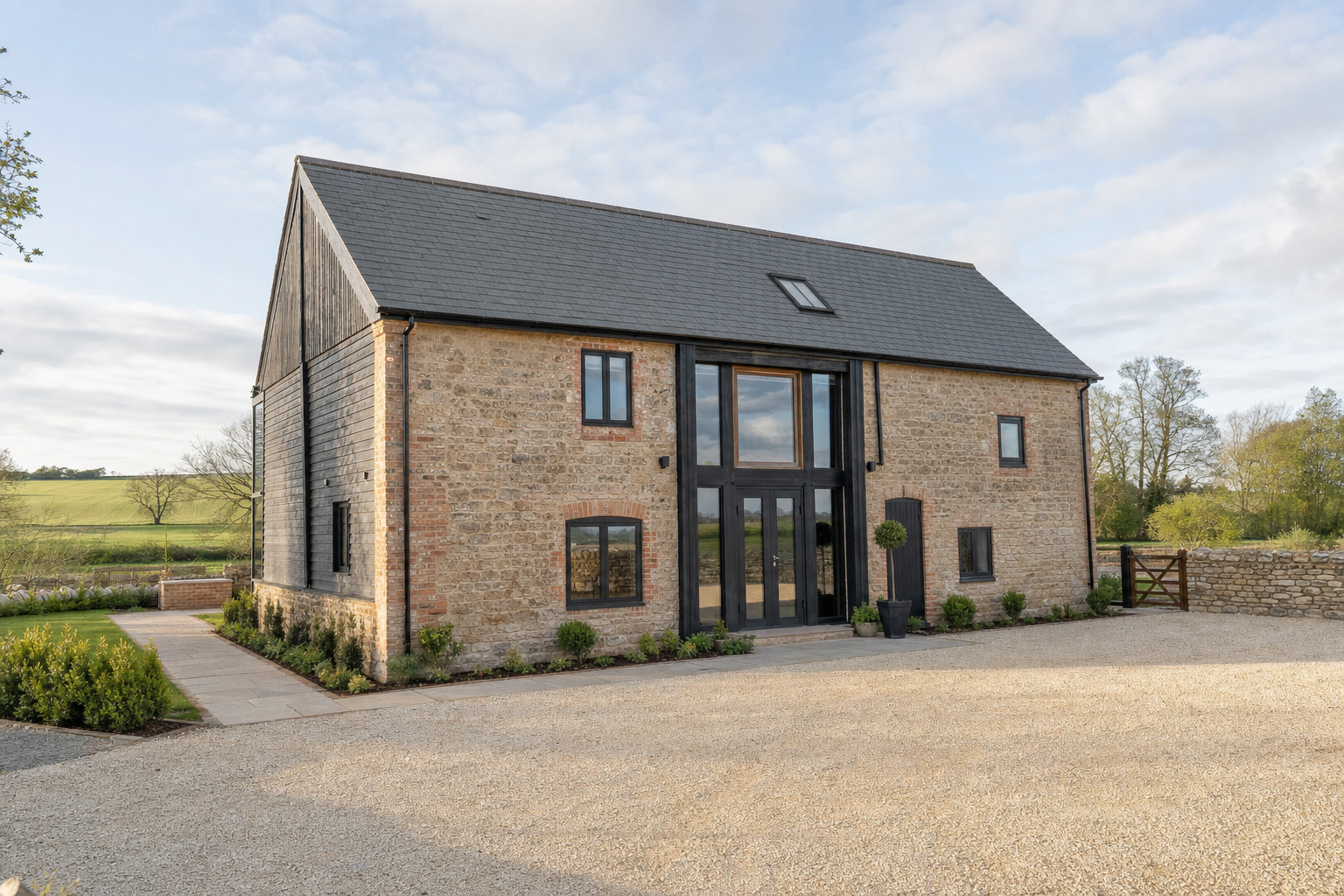 Modern barn conversion with stone walls, large glass entrance, and surrounding countryside under a partly cloudy sky.
