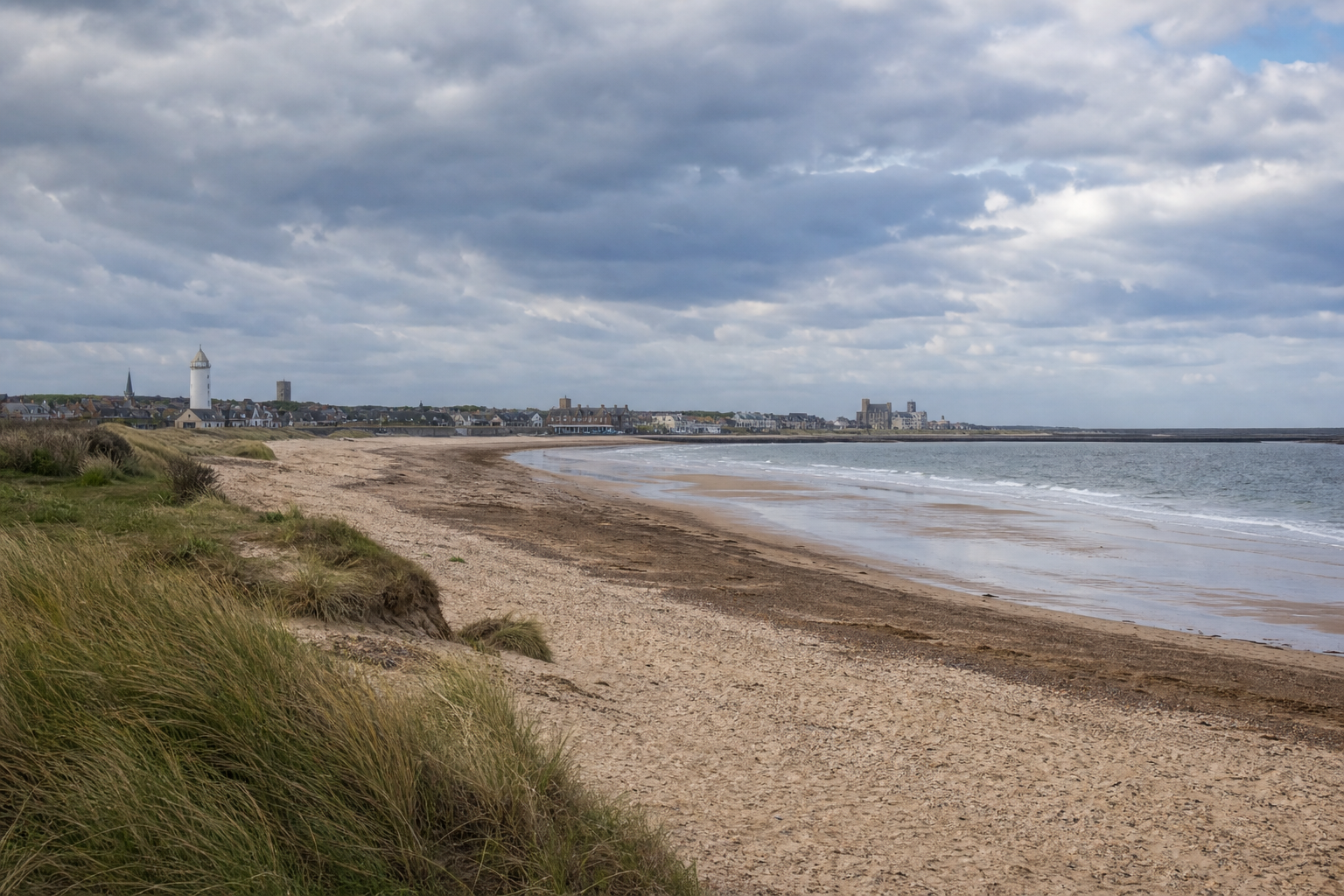 A sandy beach with grassy dunes leads to a distant coastal town under a cloudy sky.
