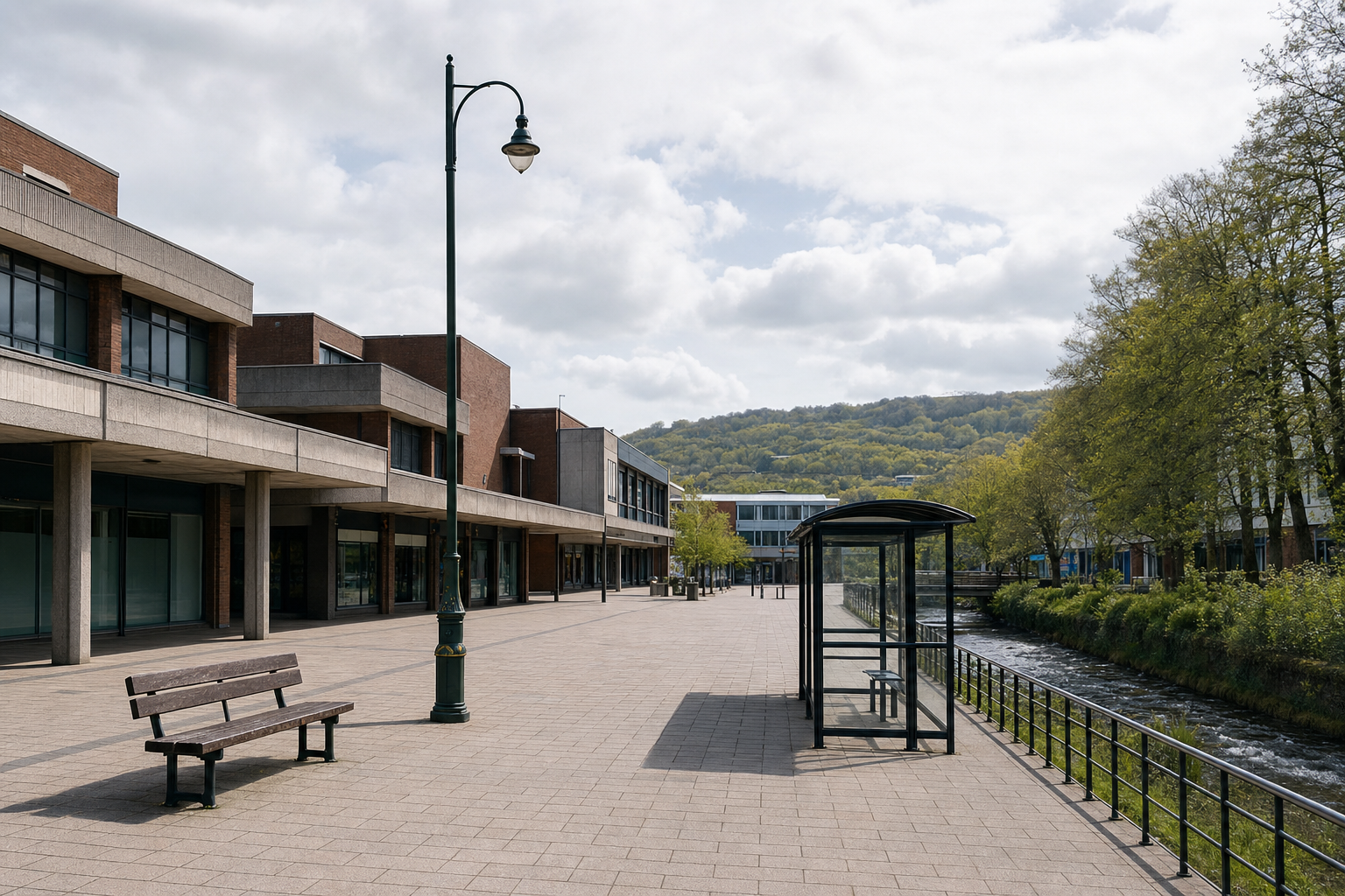An empty street with a bench and bus stop by a river, flanked by modern buildings and hills in the background.