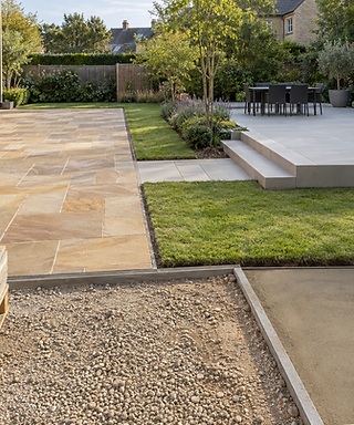 A modern garden with stone patios, green lawn, outdoor dining area, and paving in progress.