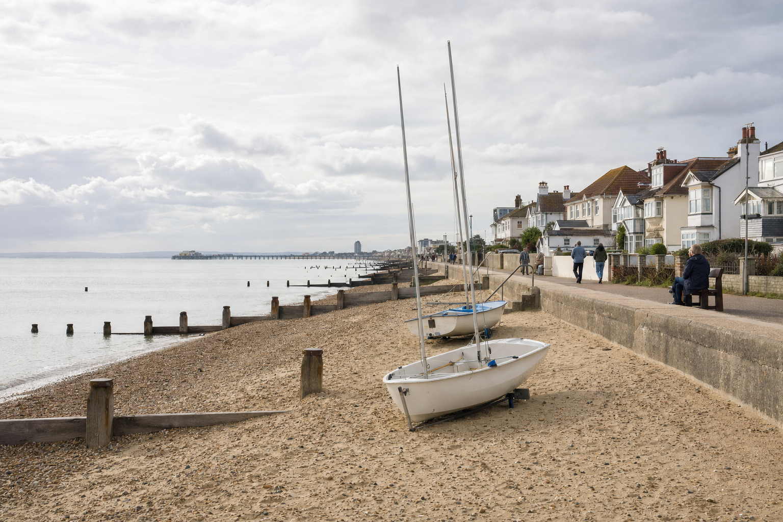 Sailboats rest on a pebbled beach beside a row of seaside houses under a cloudy sky.